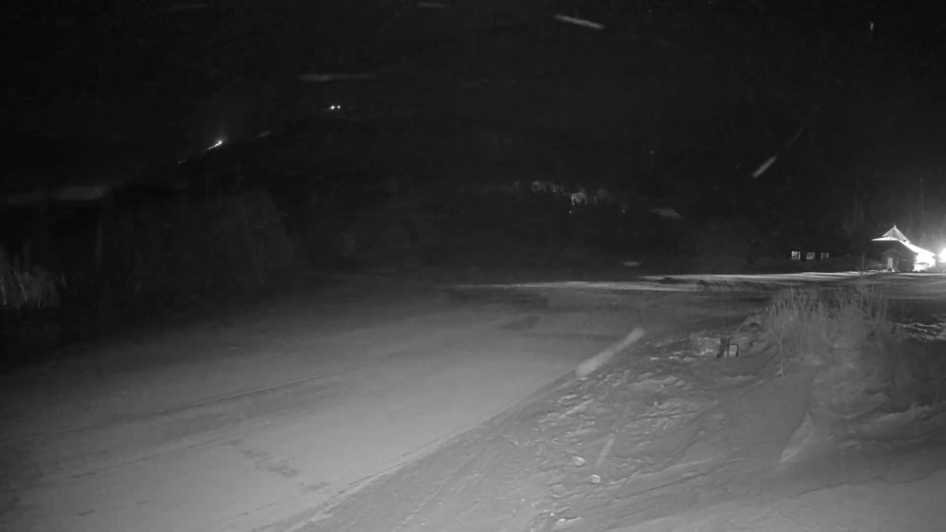 A snow-covered, dark road at night with a building faintly visible in the distance under falling snow.
