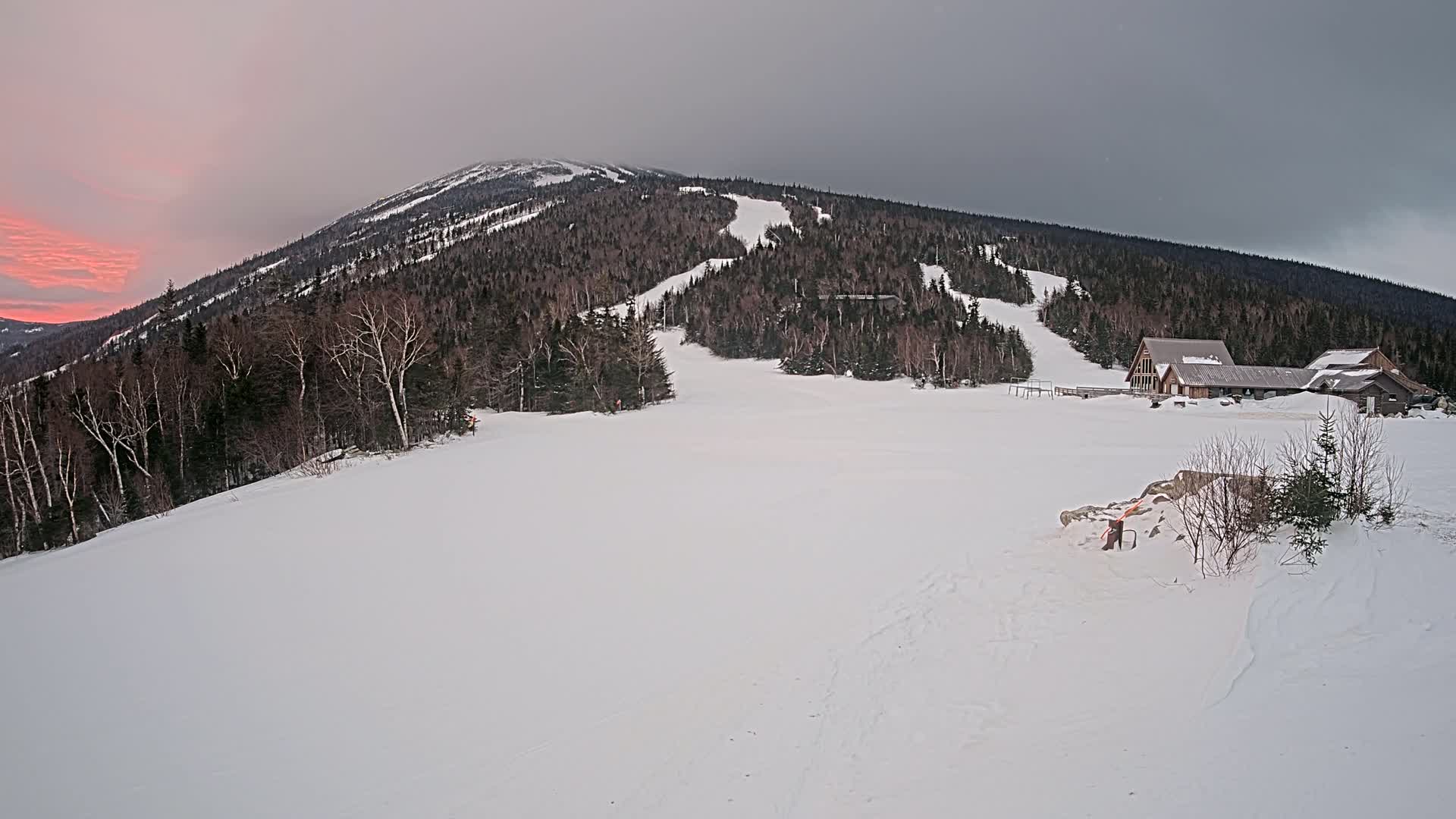 A snow-covered mountain slope with a ski lodge at its base, under a cloudy sky with a pinkish sunset.