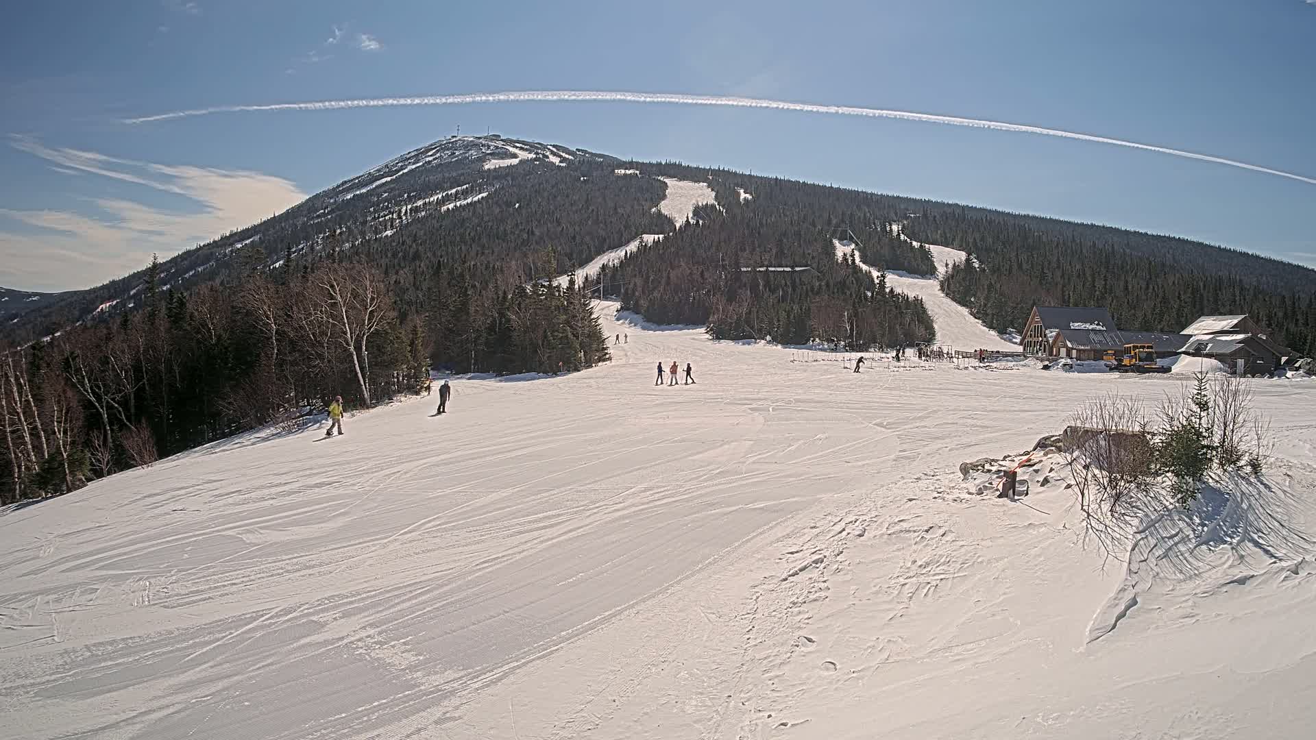A snow-covered ski slope with a few skiers and snowboarders, a ski lodge, and a mountain in the background under a clear blue sky.
