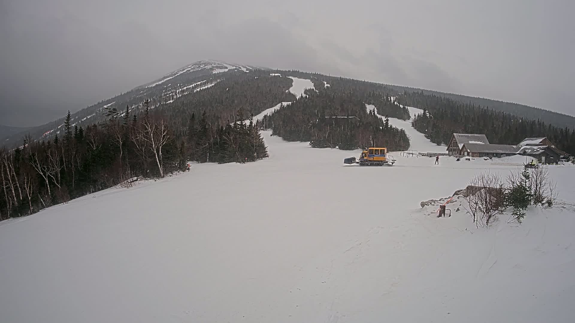 A snowcat grooms a snow-covered ski slope beneath an overcast sky, with a mountain in the background and a small building near the bottom of the slope.