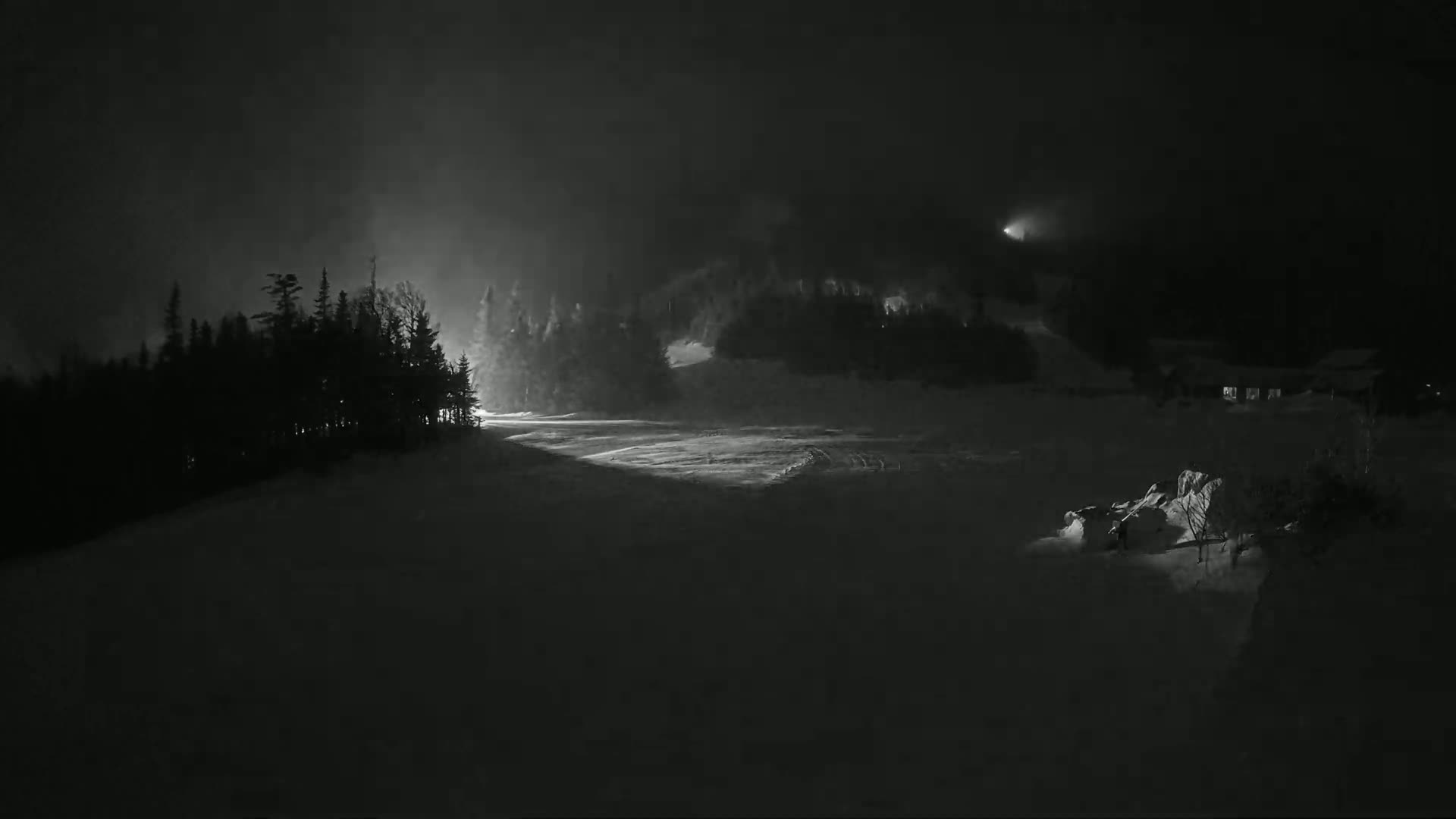 A snow-covered landscape at night, illuminated by faint lights, shows dark silhouettes of trees and buildings under hazy, dark conditions.