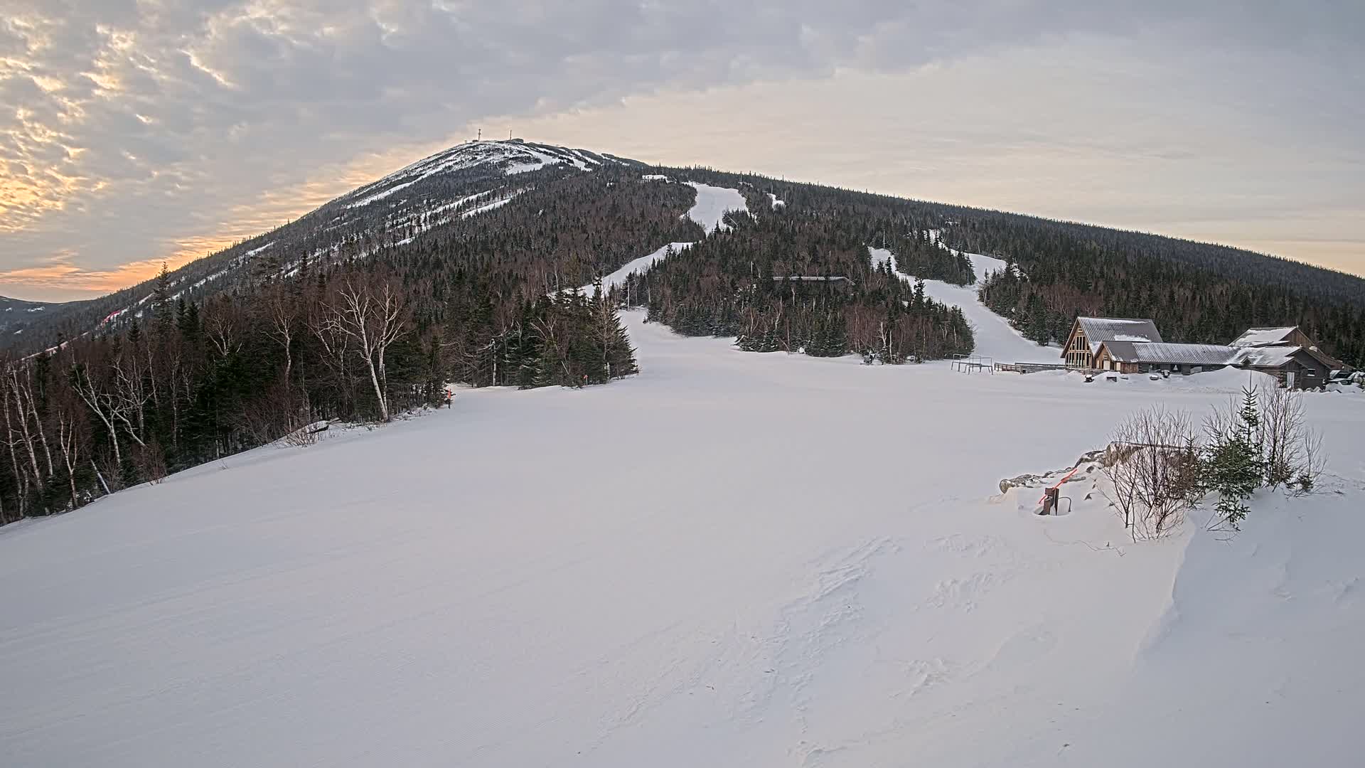 A snow-covered ski slope at sunset, with a mountain in the background and a few buildings near the base.