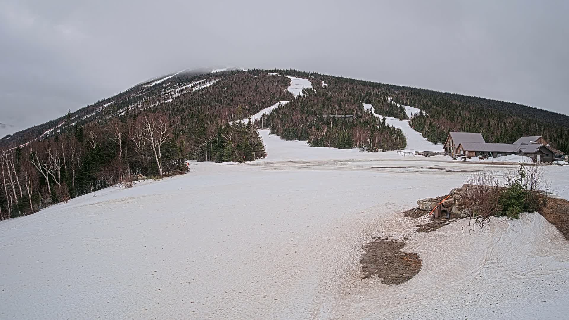 A snow-covered mountain slope with a ski lodge and several ski runs visible under an overcast sky.