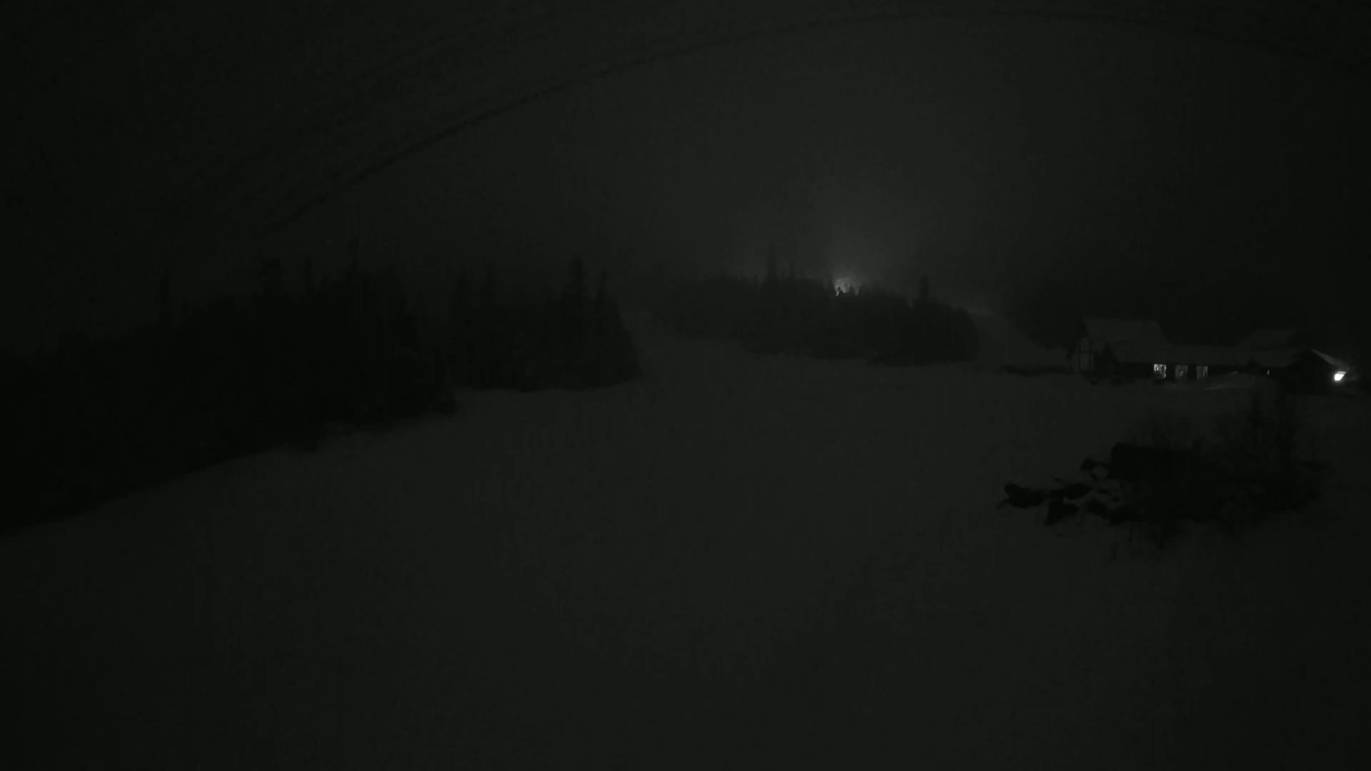 A dimly lit snowy landscape at night, shrouded in fog, shows a dark building with illuminated windows in the lower right corner.