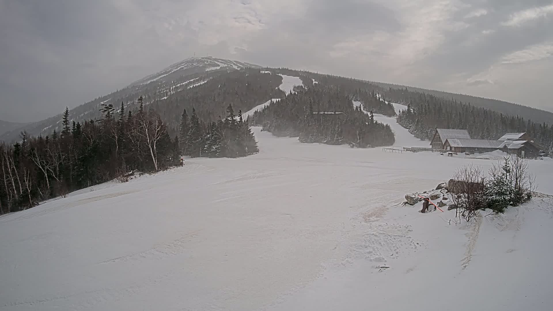A snow-covered ski slope at the base of a large mountain under overcast skies with light snow falling.