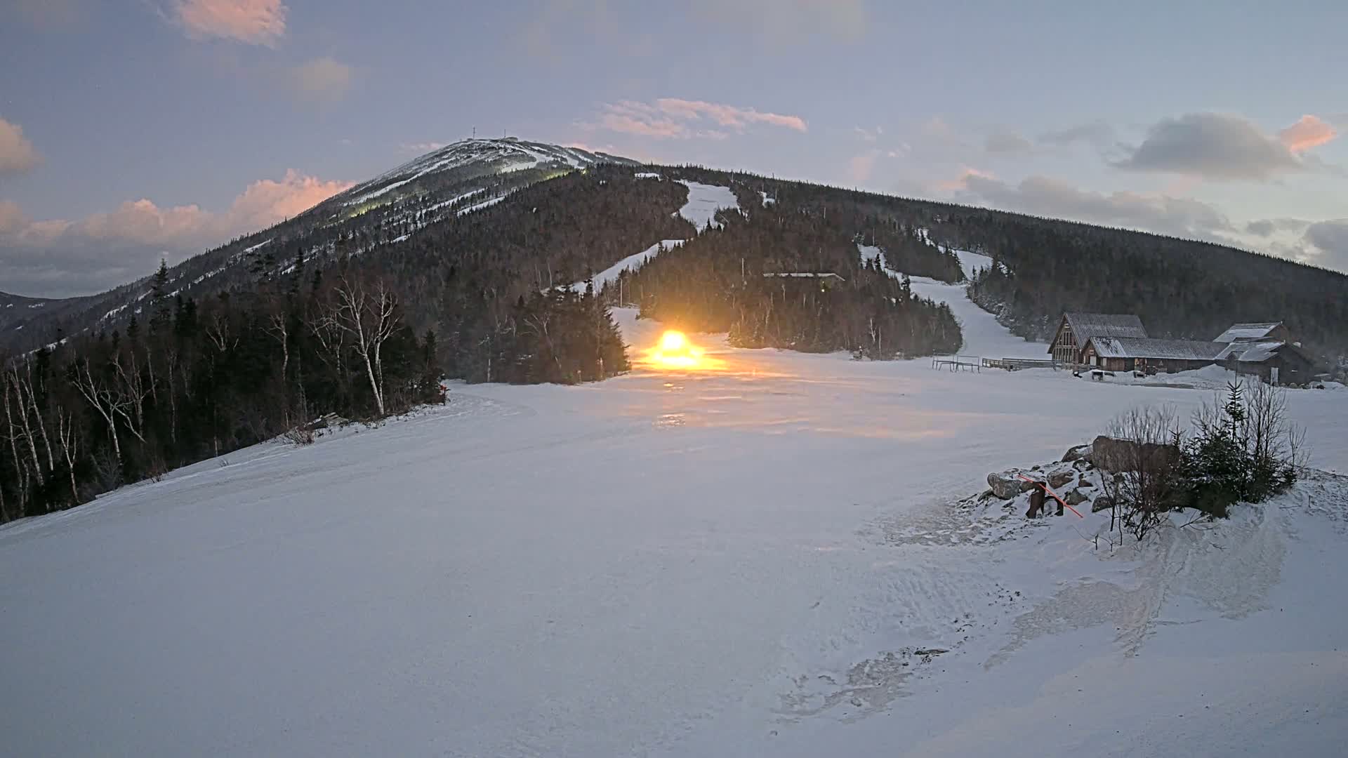 A snow-covered ski slope at dusk, with a bright light emanating from a snowcat near the base of a mountain.