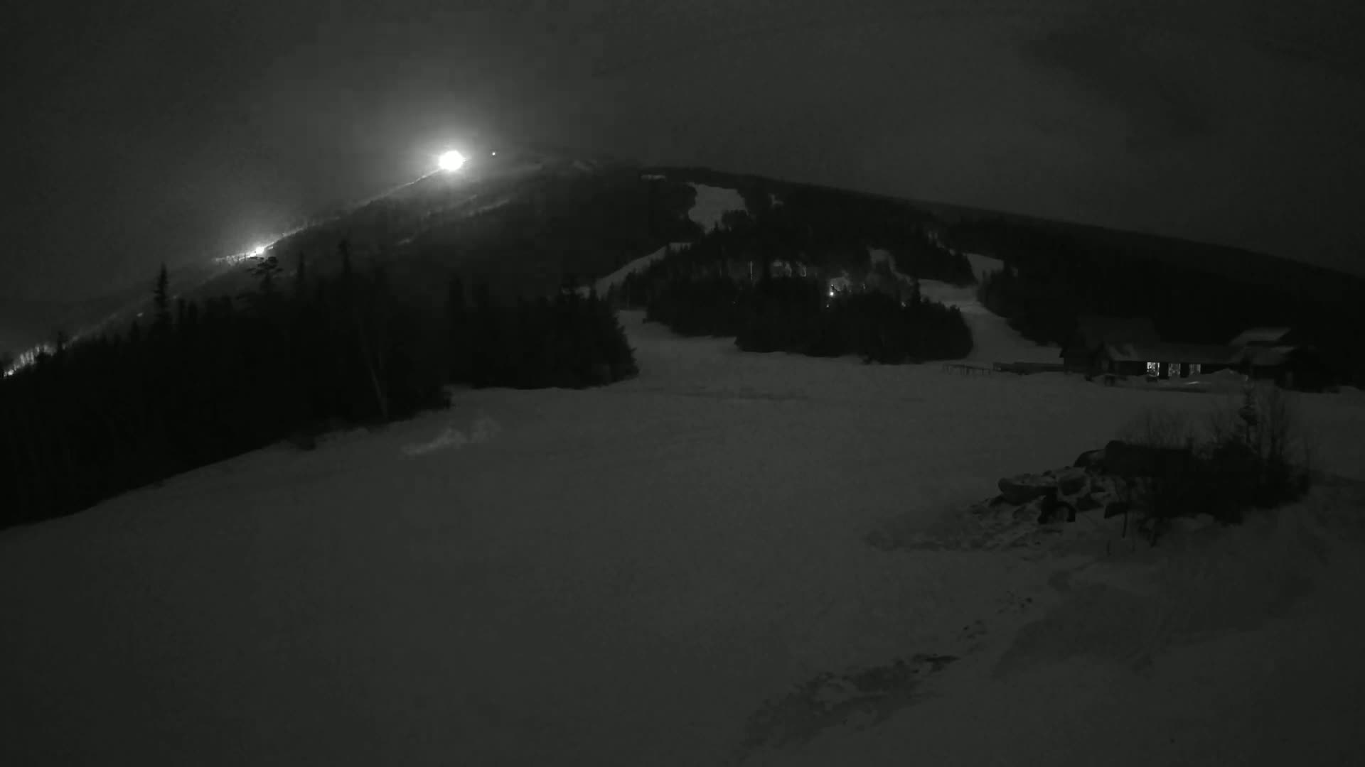 A snow-covered hill at night, illuminated by lights on the slope and at the base of the hill, with dark evergreen trees.