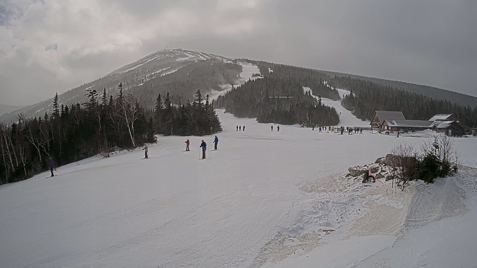 A snow-covered ski slope with several skiers and a mountain in the background under an overcast sky.