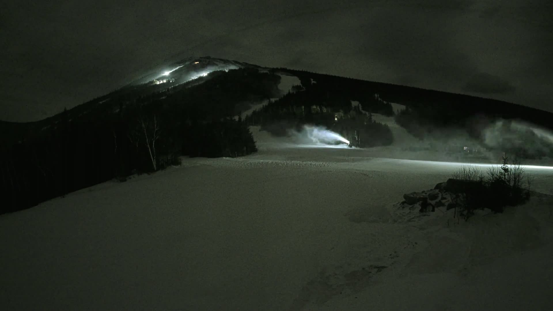 Snowmaking machines illuminate a snow-covered ski slope at night under a dark, cloudy sky.