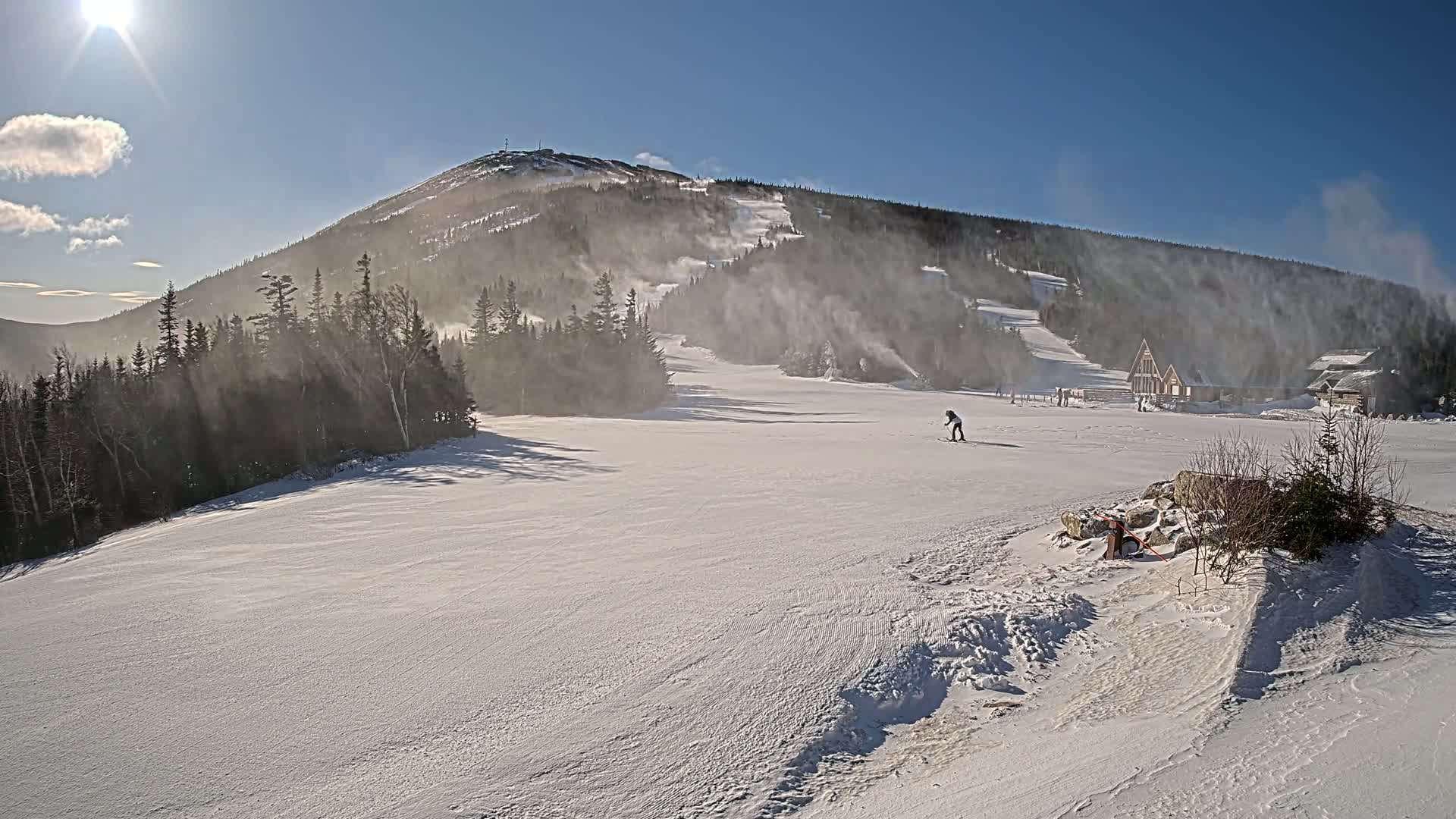 A sunny, windy day on a snow-covered ski slope with a mountain in the background, snowmaking machines are operating, and a few skiers are visible.