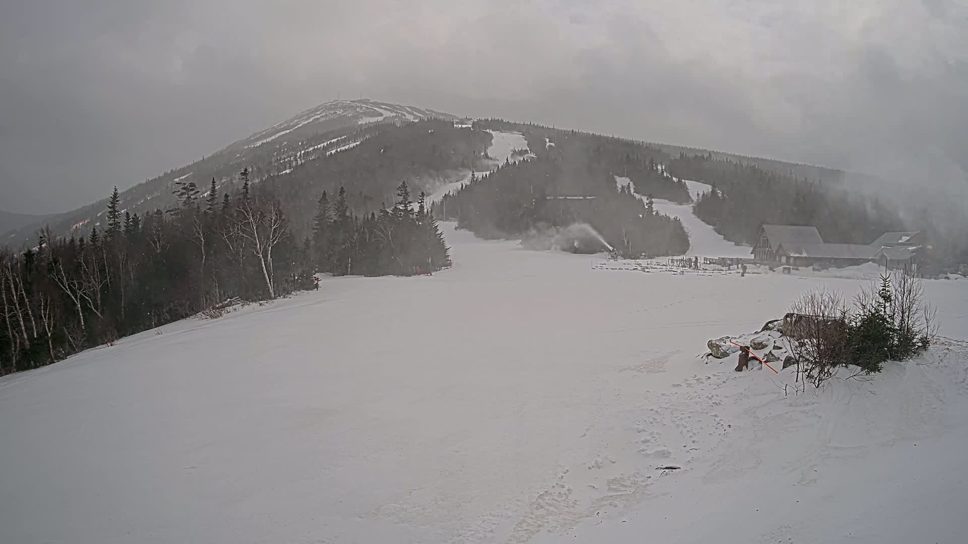 A snow-covered ski slope with a snowmaking machine operating is visible beneath a cloudy, snowy sky, with a mountain in the background.