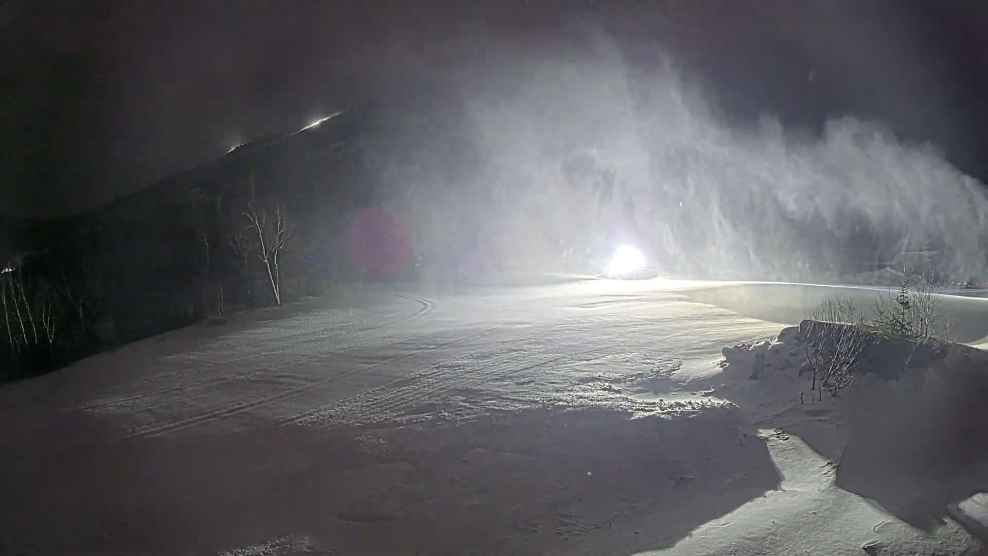 A snow-covered landscape at night, with blowing snow obscuring a vehicle's headlights in the distance.