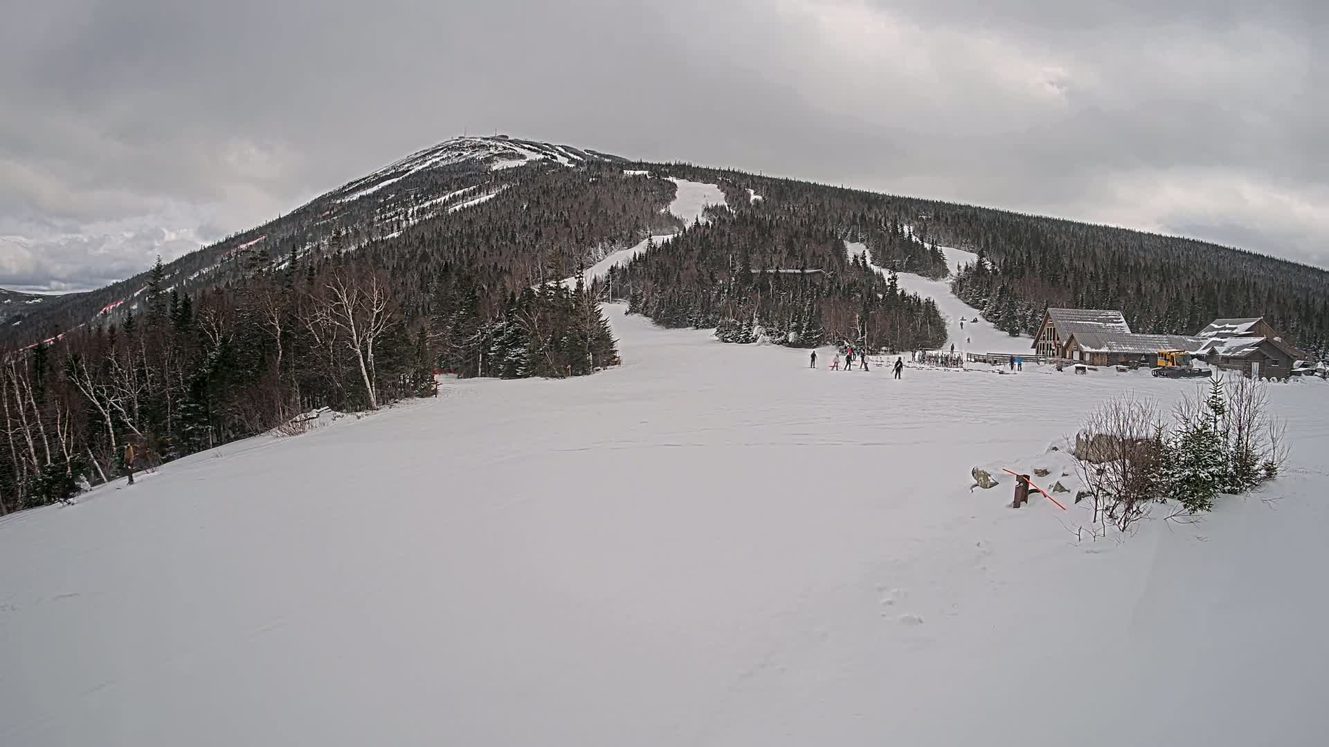 A snow-covered ski slope leads to a mountaintop under a cloudy sky, with a few skiers visible and buildings at the base.