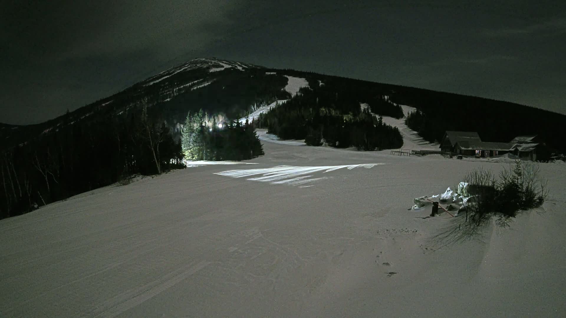 A snow-covered ski slope at night, illuminated by artificial lights, is situated below a dark, snow-capped mountain, with a small building visible in the distance.