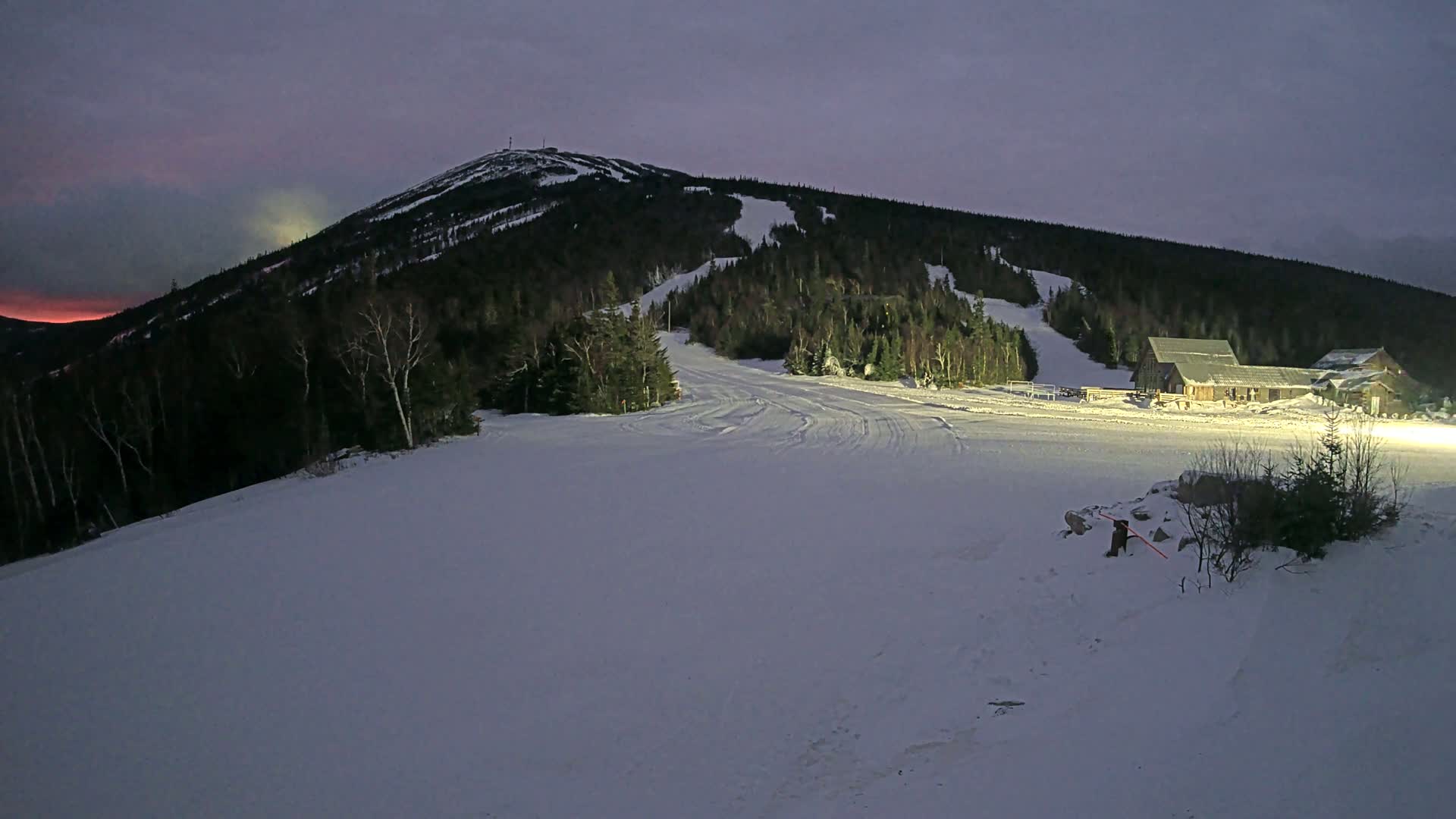 A snow-covered ski slope at dusk, with a mountain in the background and a building illuminated at the base of the hill.