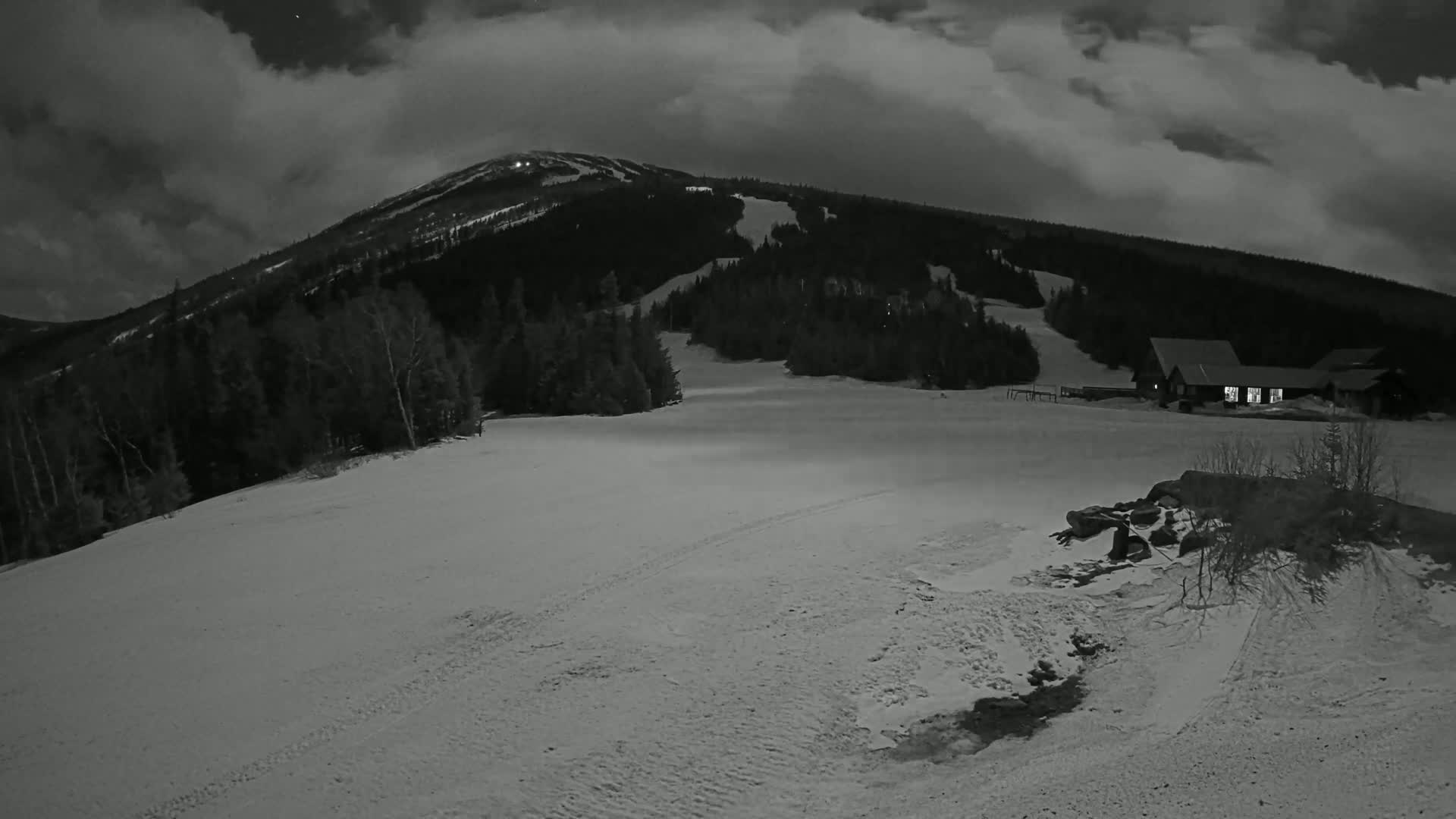 A snow-covered ski slope at night under a cloudy sky, with a building and a mountain in the background.