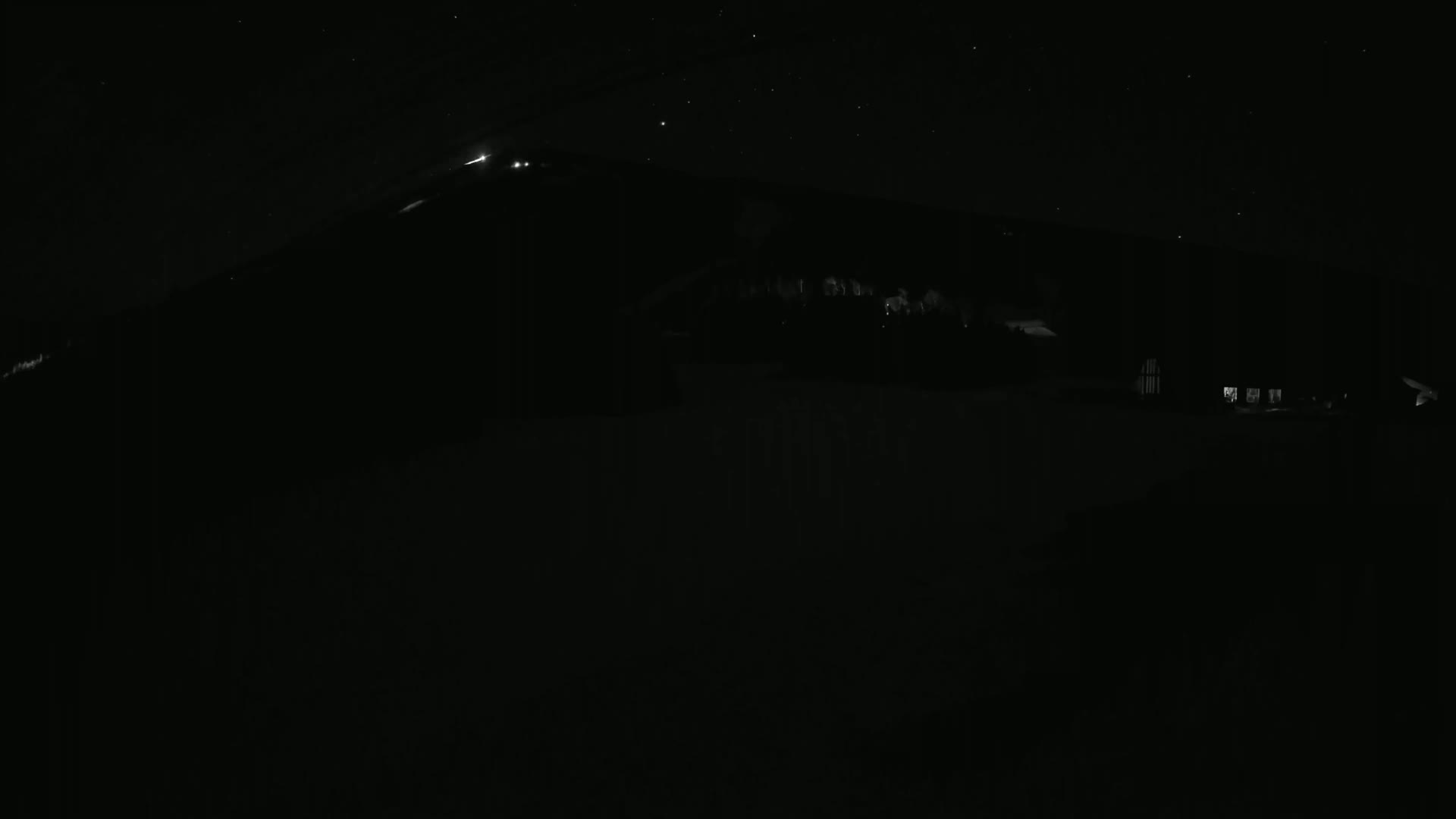 A dark, starlit night scene shows a silhouetted hill with a few faint lights from buildings at its base.