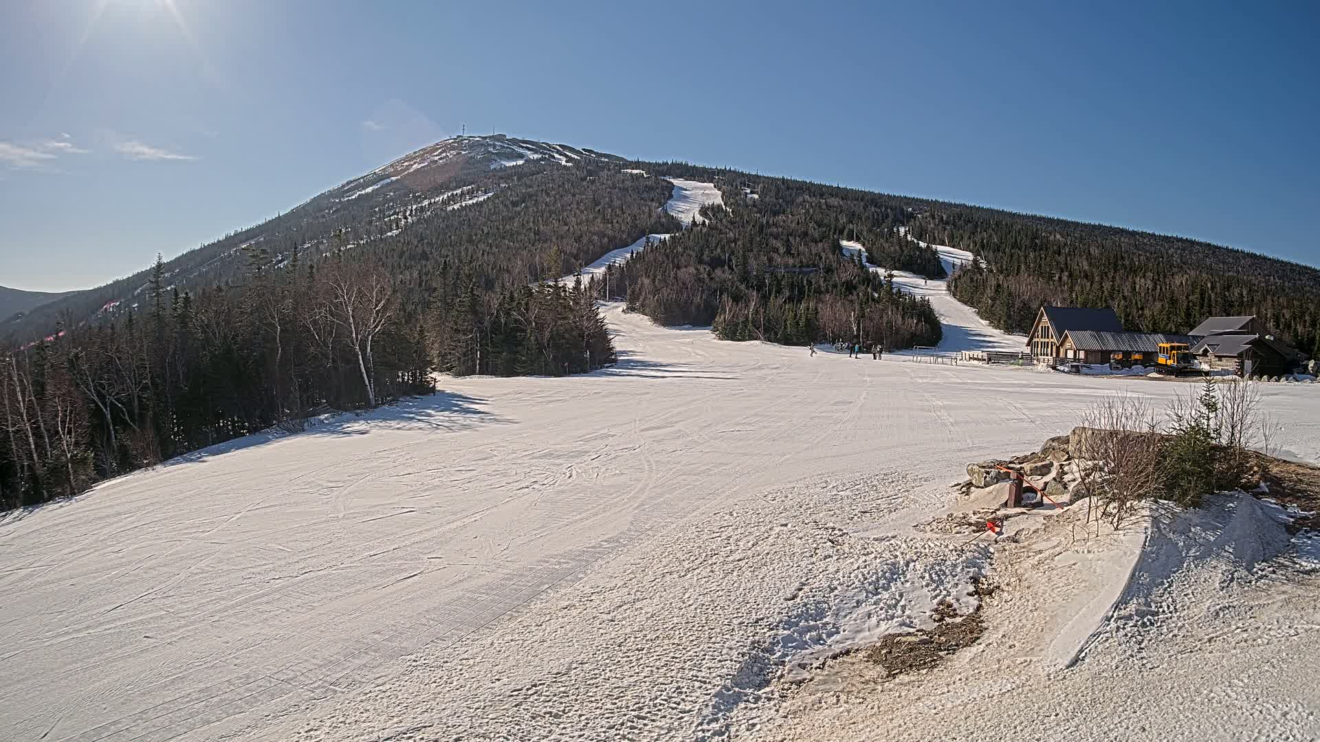 A snow-covered ski slope leads to a mountain peak under a bright, sunny sky.