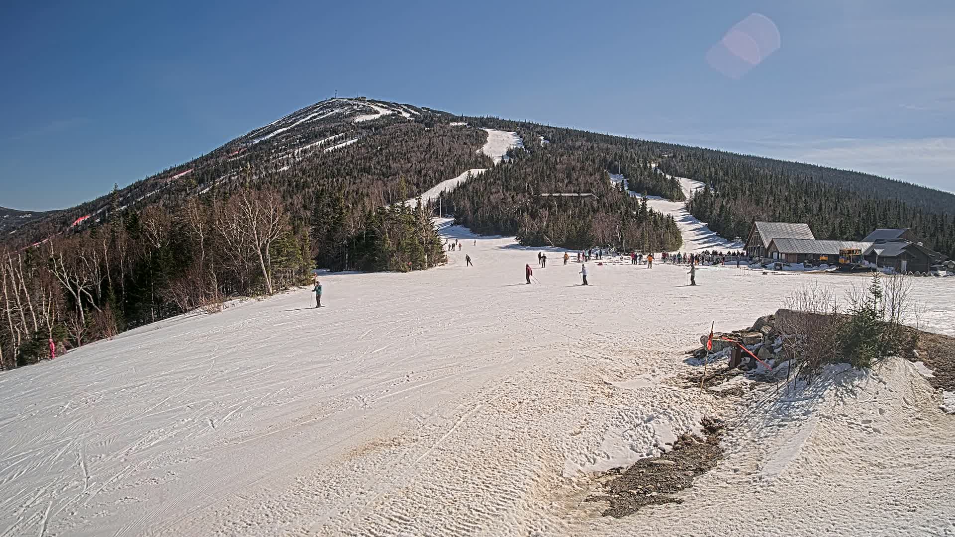 A snow-covered mountain slope with skiers and snowboarders, a ski lodge, and a clear blue sky.