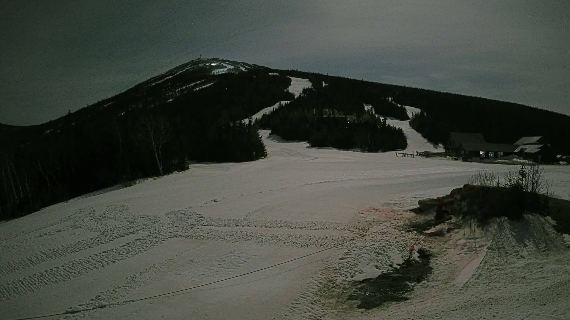 A snow-covered ski slope at night, with a dark mountain in the background and a few buildings near the bottom.