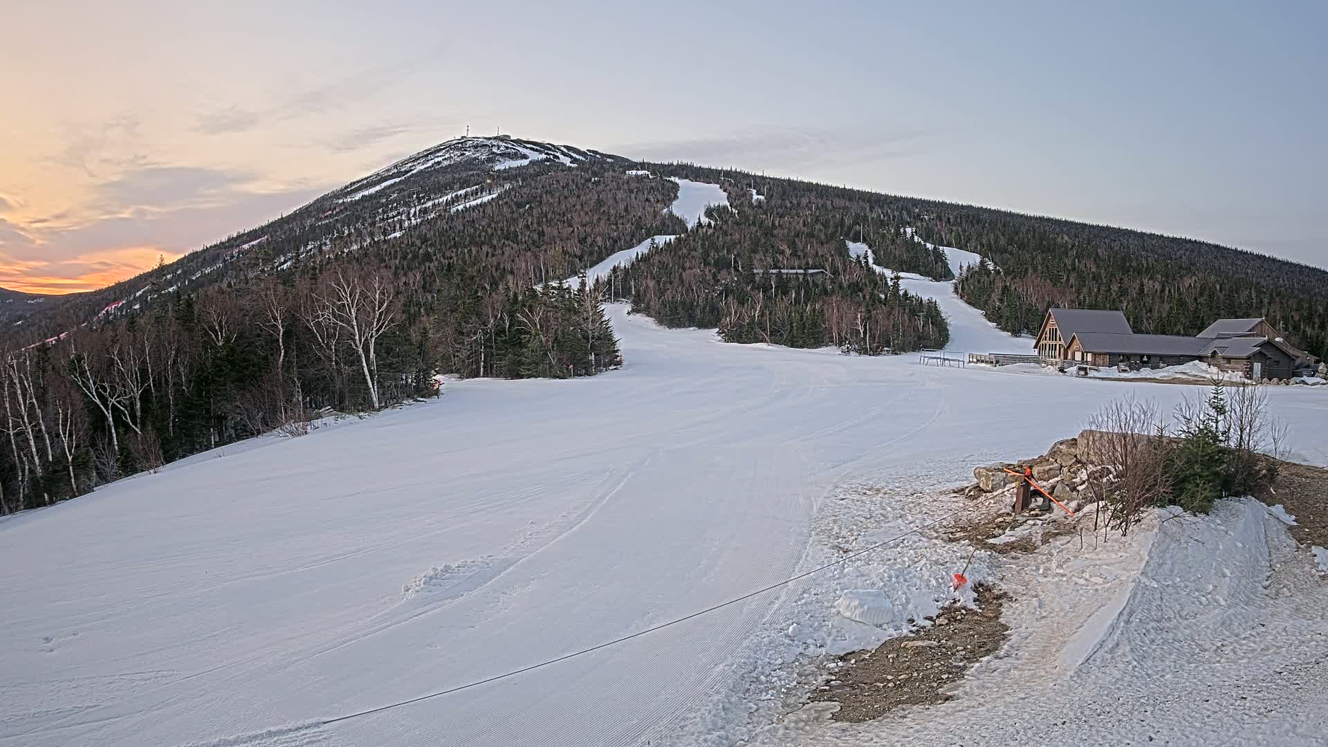A snow-covered ski slope at sunset, with a mountain in the background and a lodge near the bottom.