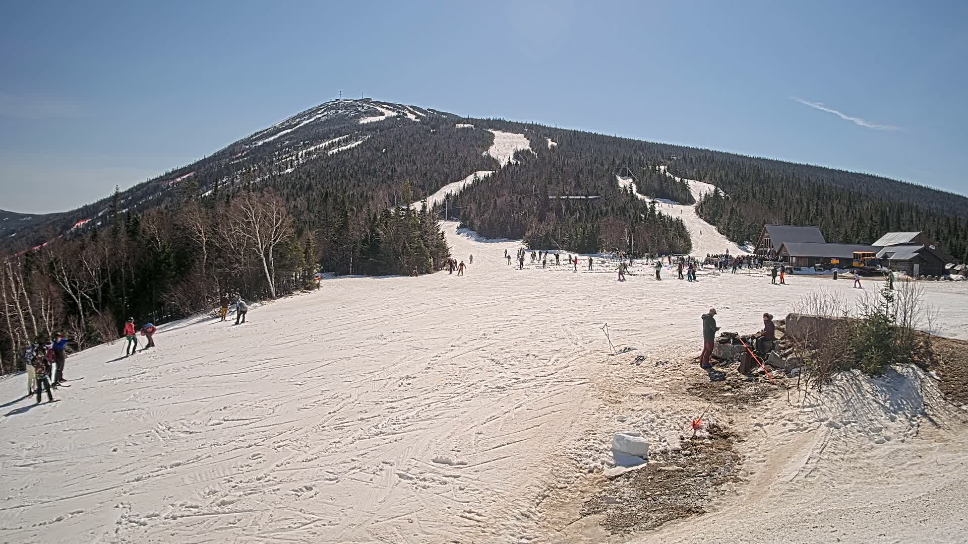 A snow-covered ski slope with scattered skiers and snowboarders under a clear blue sky, with a large mountain in the background.