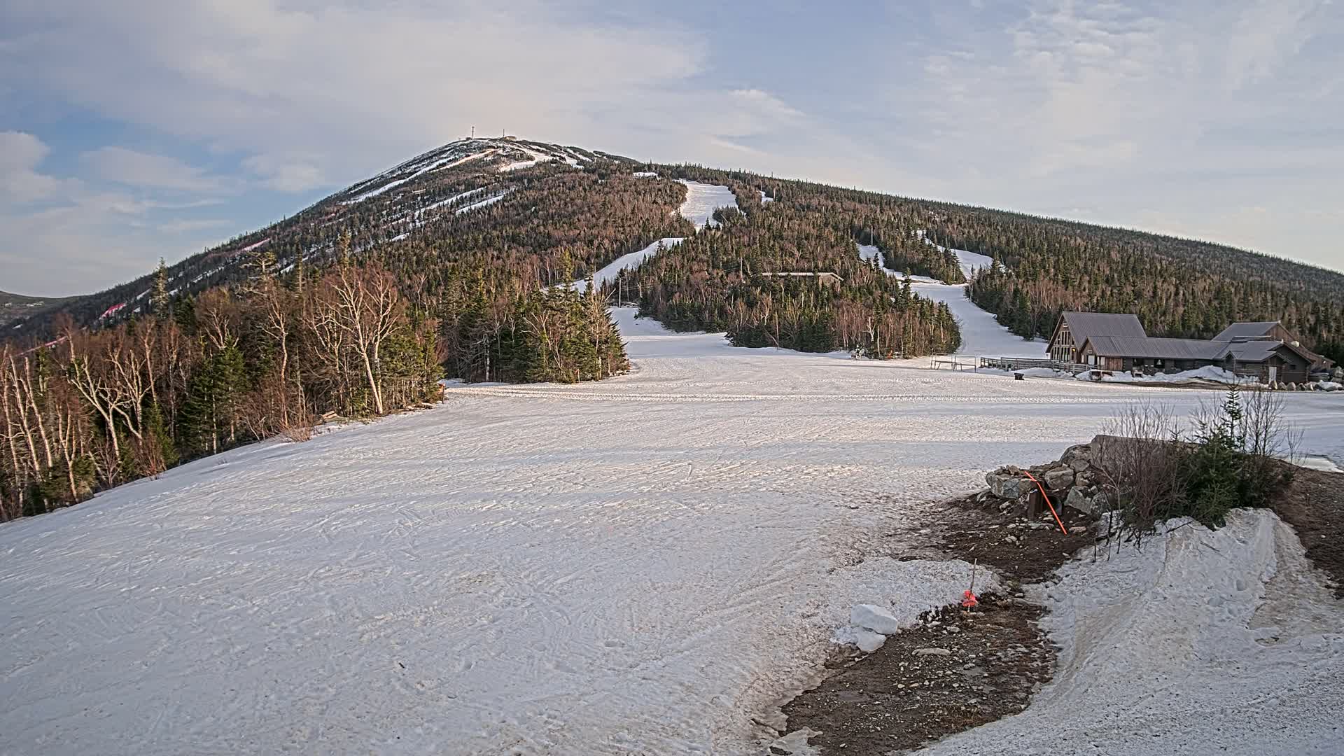 A snow-covered ski slope at the base of a mountain with a few buildings, under a partly cloudy sky.