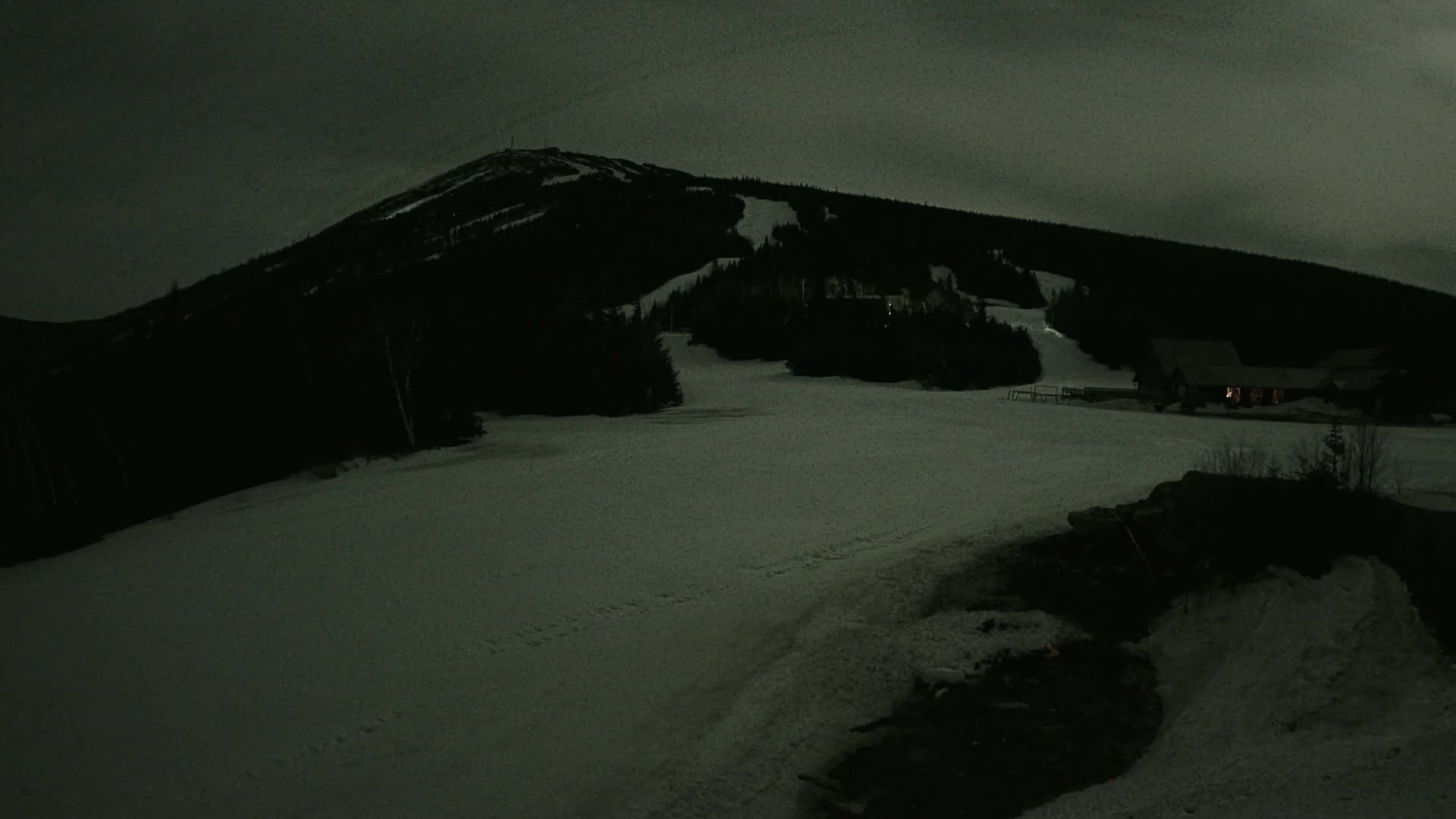 A snow-covered ski slope at night under a dark sky, with a mountain in the background and a few lights visible near the bottom.