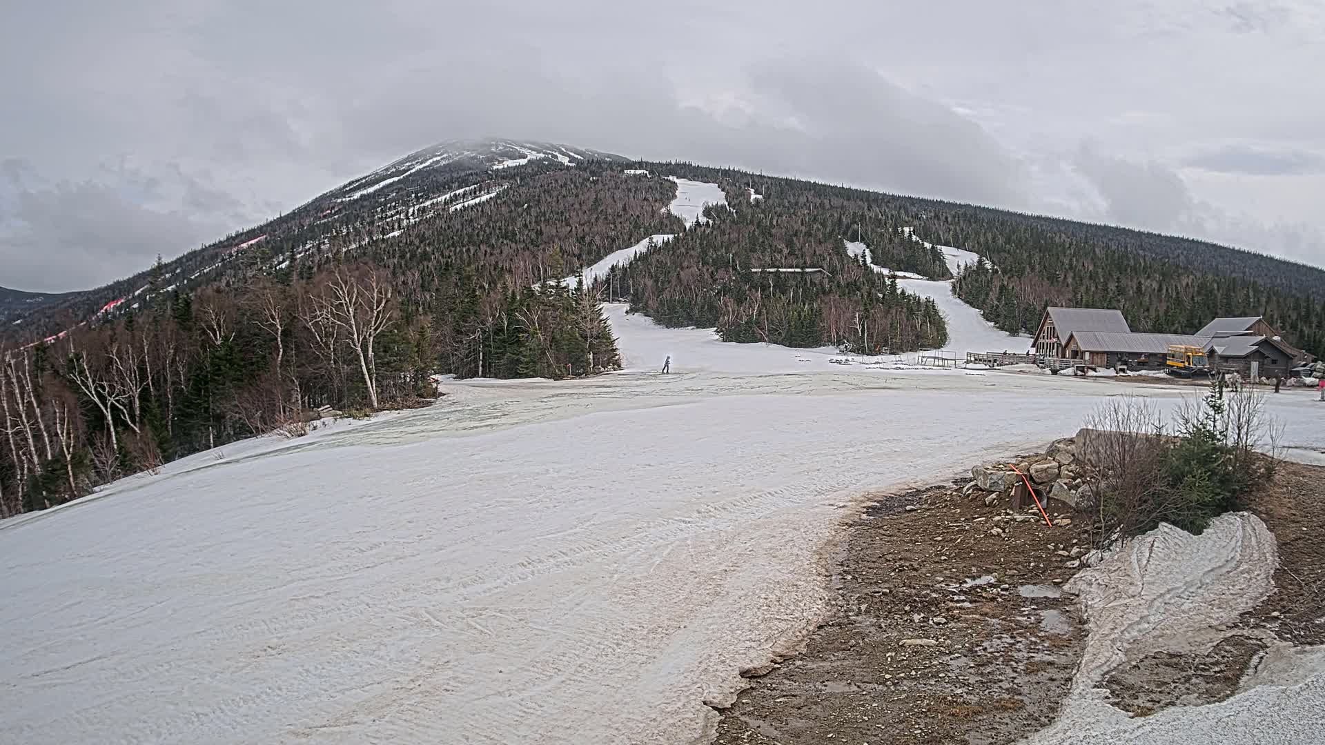 A snow-covered ski slope at the base of a forested mountain on an overcast day.