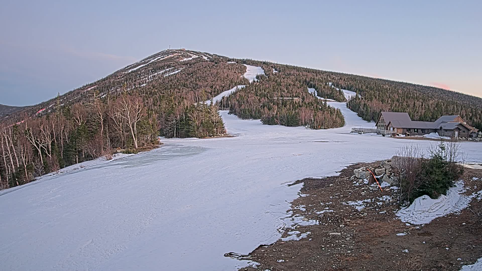 A snow-covered ski slope at the base of a mountain with a building at the bottom, under a twilight sky.