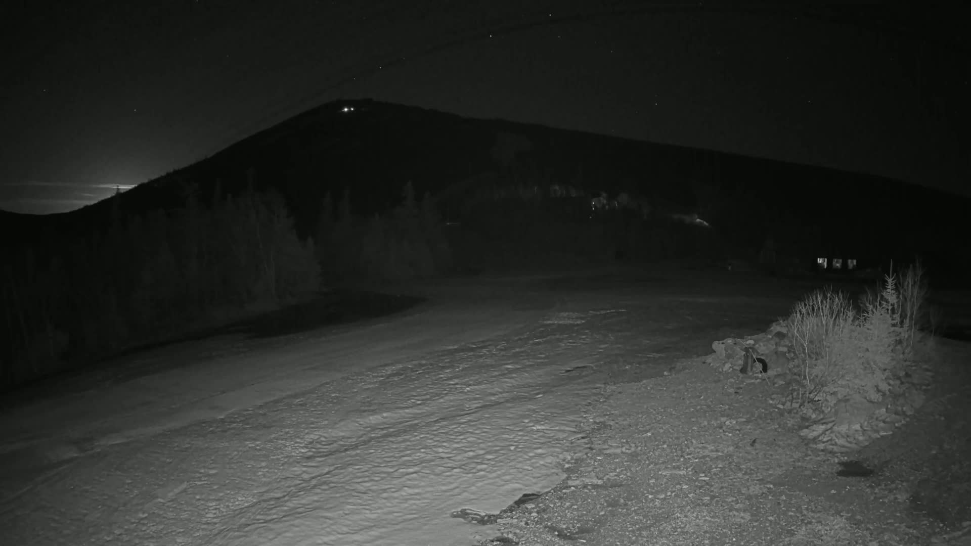 A snow-covered ground stretches out before a dark, snow-dusted mountain under a starry night sky.