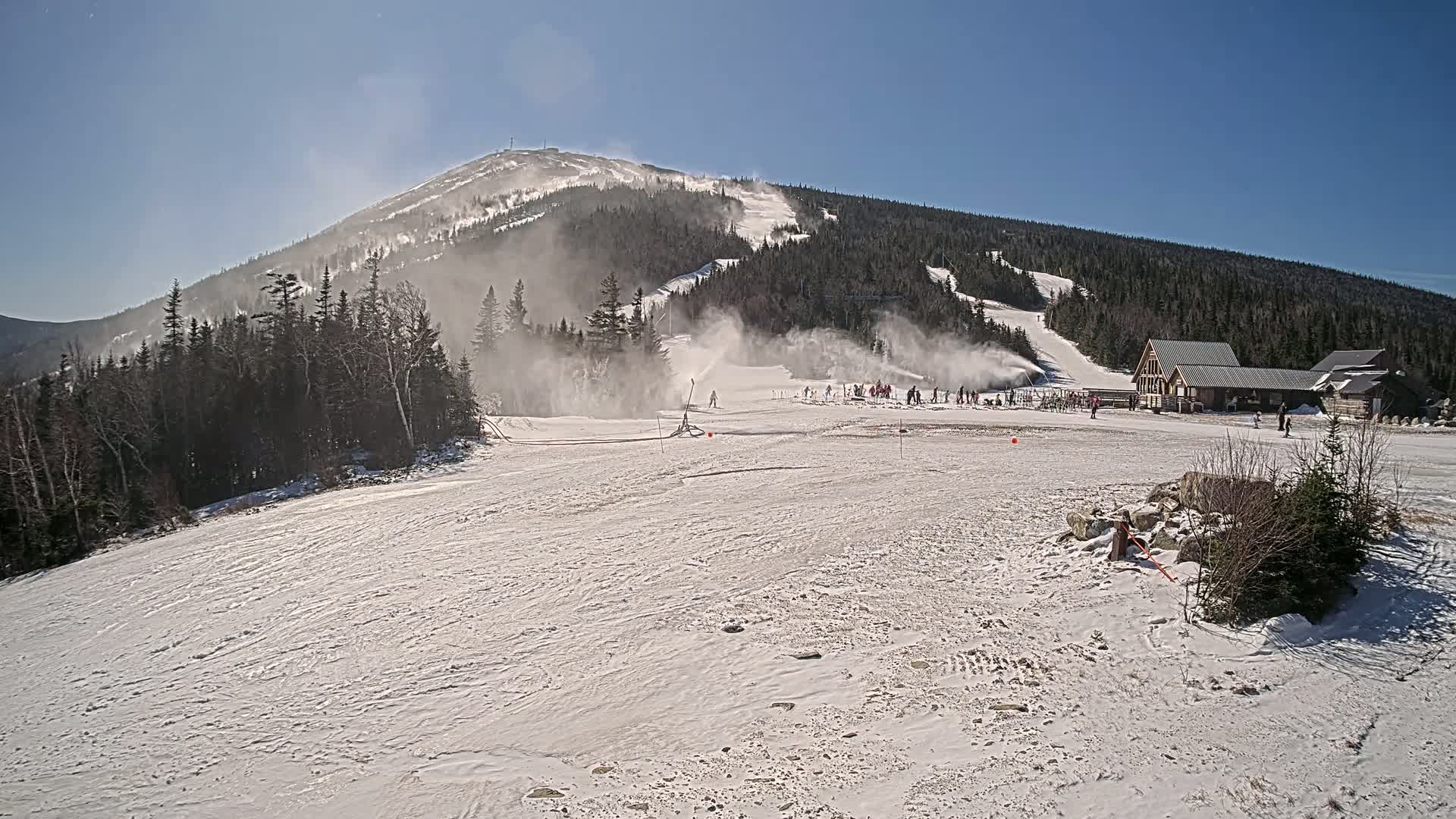 A snow-covered ski slope with snow-making machines operating on a sunny day, featuring a mountain in the background and a lodge at the bottom.