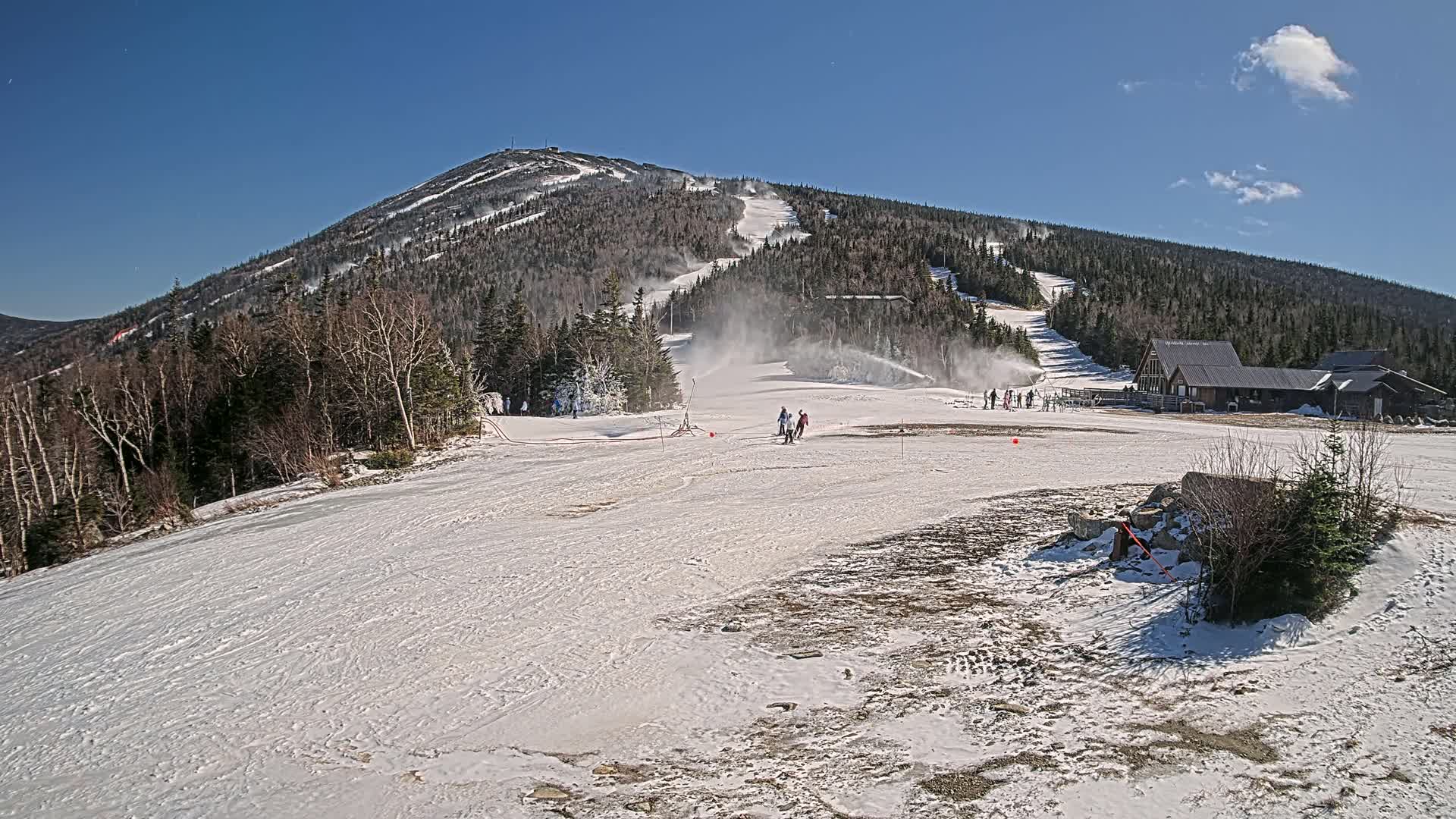 A snow-covered ski slope with several skiers and snowmaking machines operating under a clear blue sky.