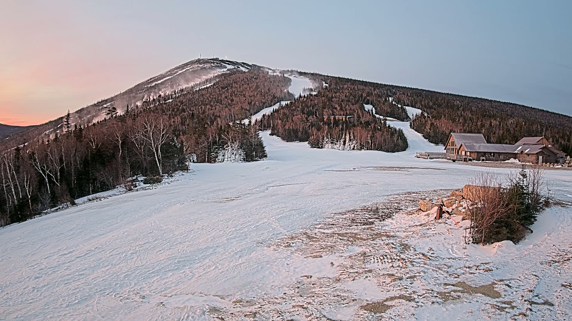 A snow-covered ski resort at sunset, featuring a mountain with ski runs, a few buildings, and some sparsely snow-covered ground in the foreground.