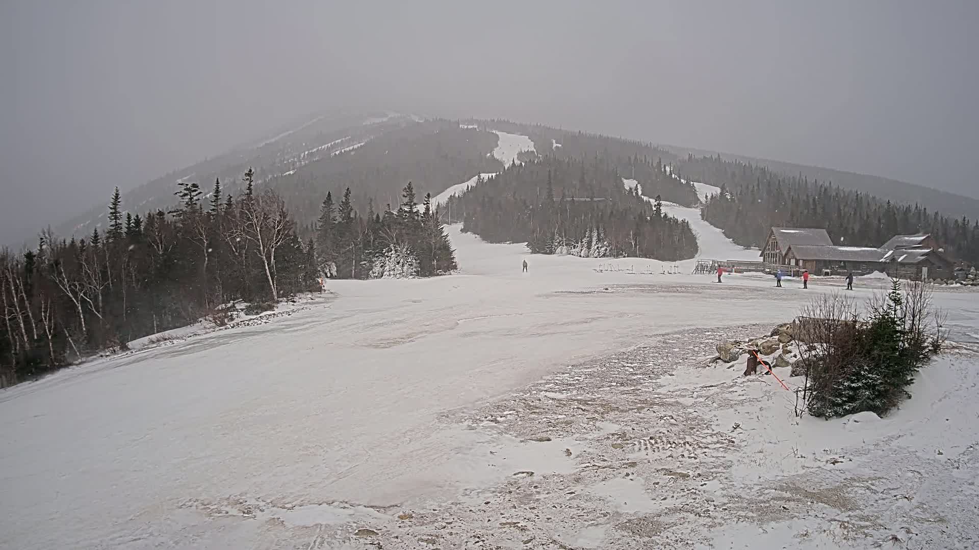 A snow-covered ski slope with a few skiers and a building in the background, under heavy snowfall.