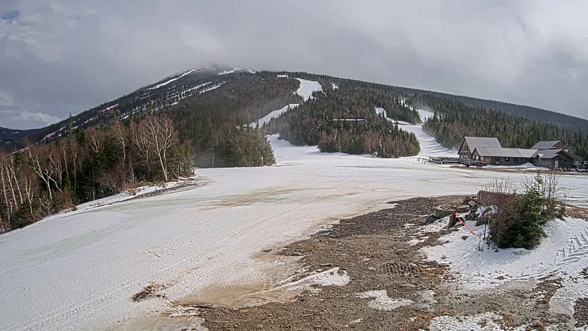 A snowy mountain ski slope with a few buildings at the base under a cloudy sky.