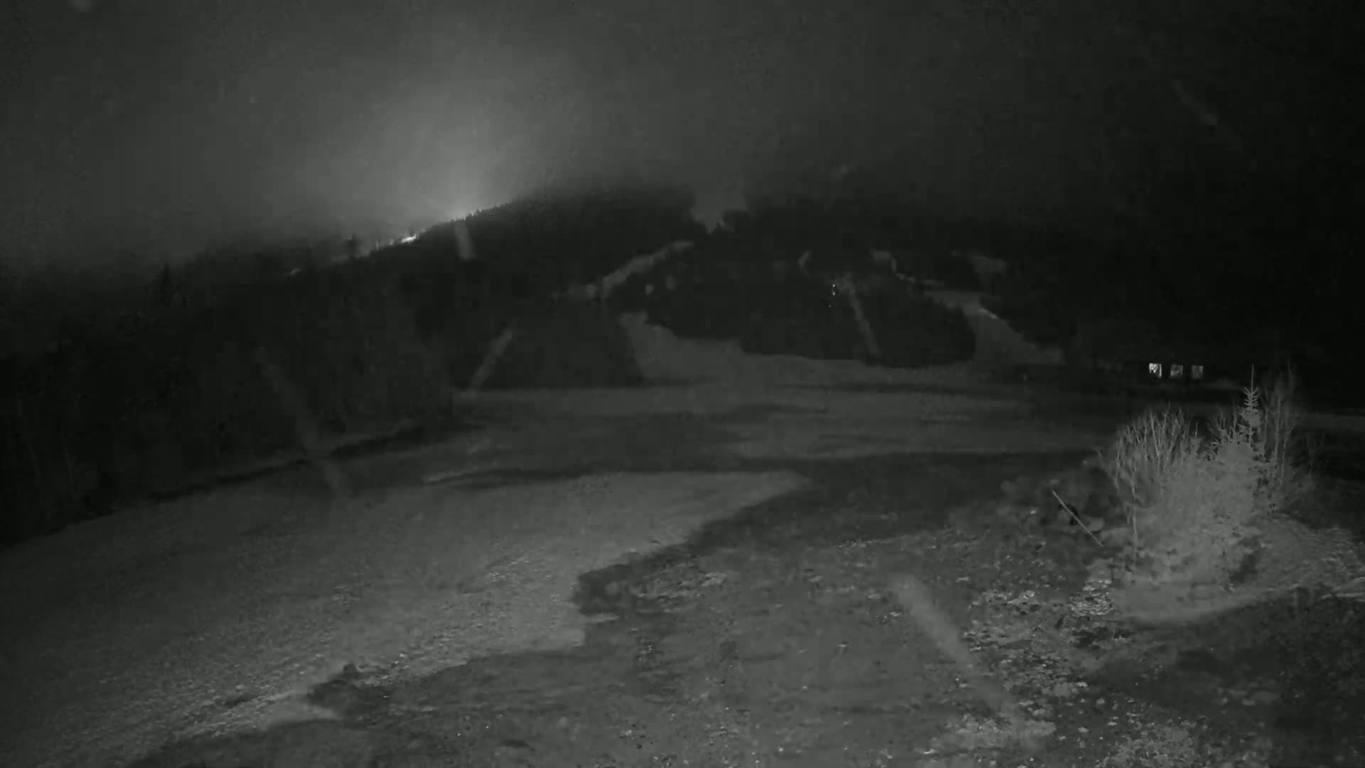 A nighttime, snowy landscape shows a sparsely vegetated area in the foreground, with a dark, snow-covered hill rising in the background under a hazy sky.