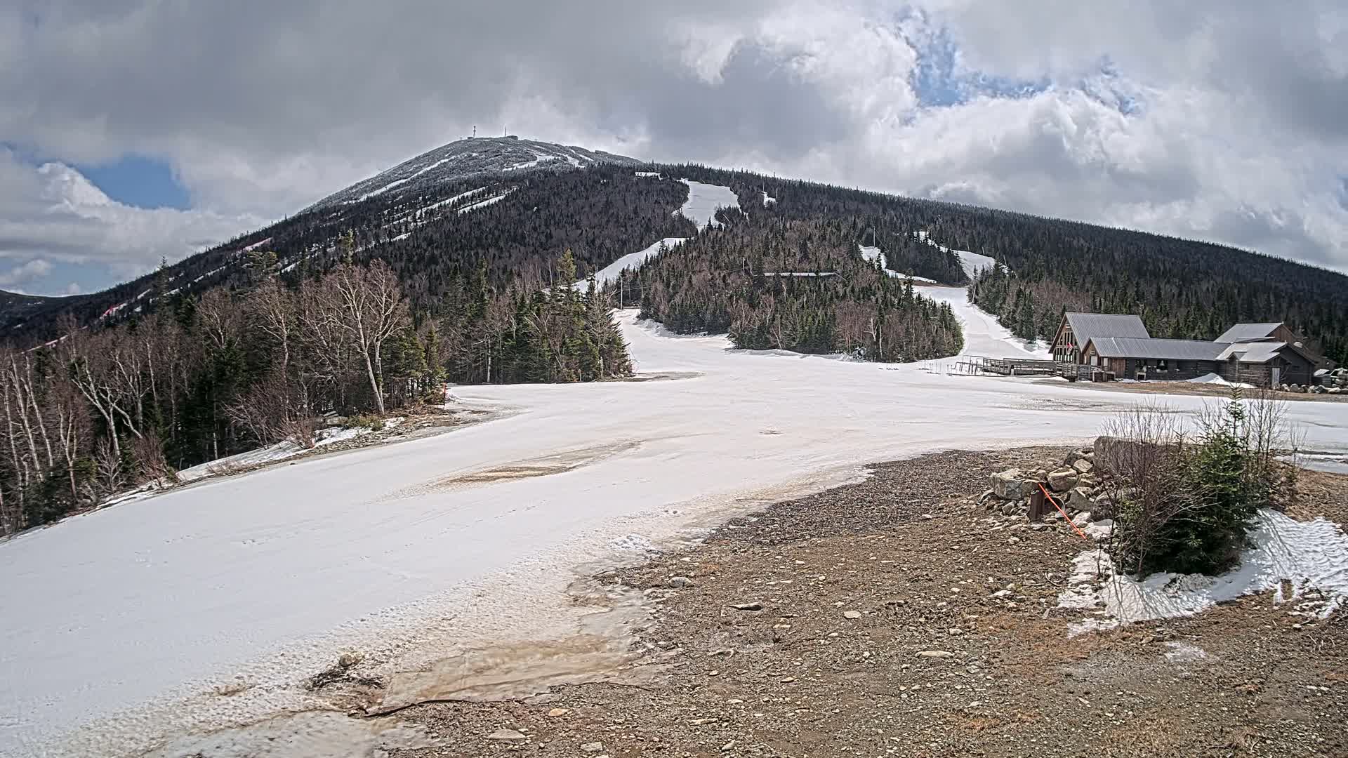 A snow-covered ski slope at the base of a mountain under a partly cloudy sky.