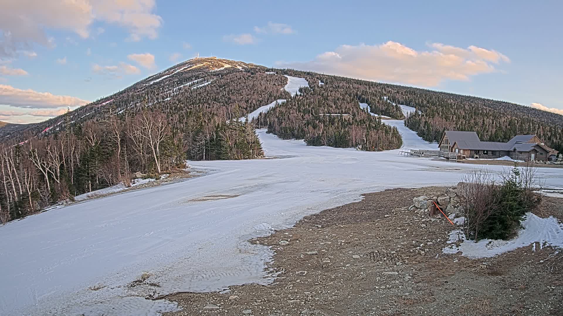 A snow-covered ski slope at the base of a mountain under a partly cloudy sky.