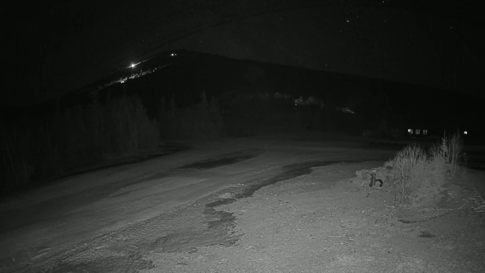 A nighttime, snow-covered, gravelly area stretches toward a dark mountain silhouetted against a starry sky.