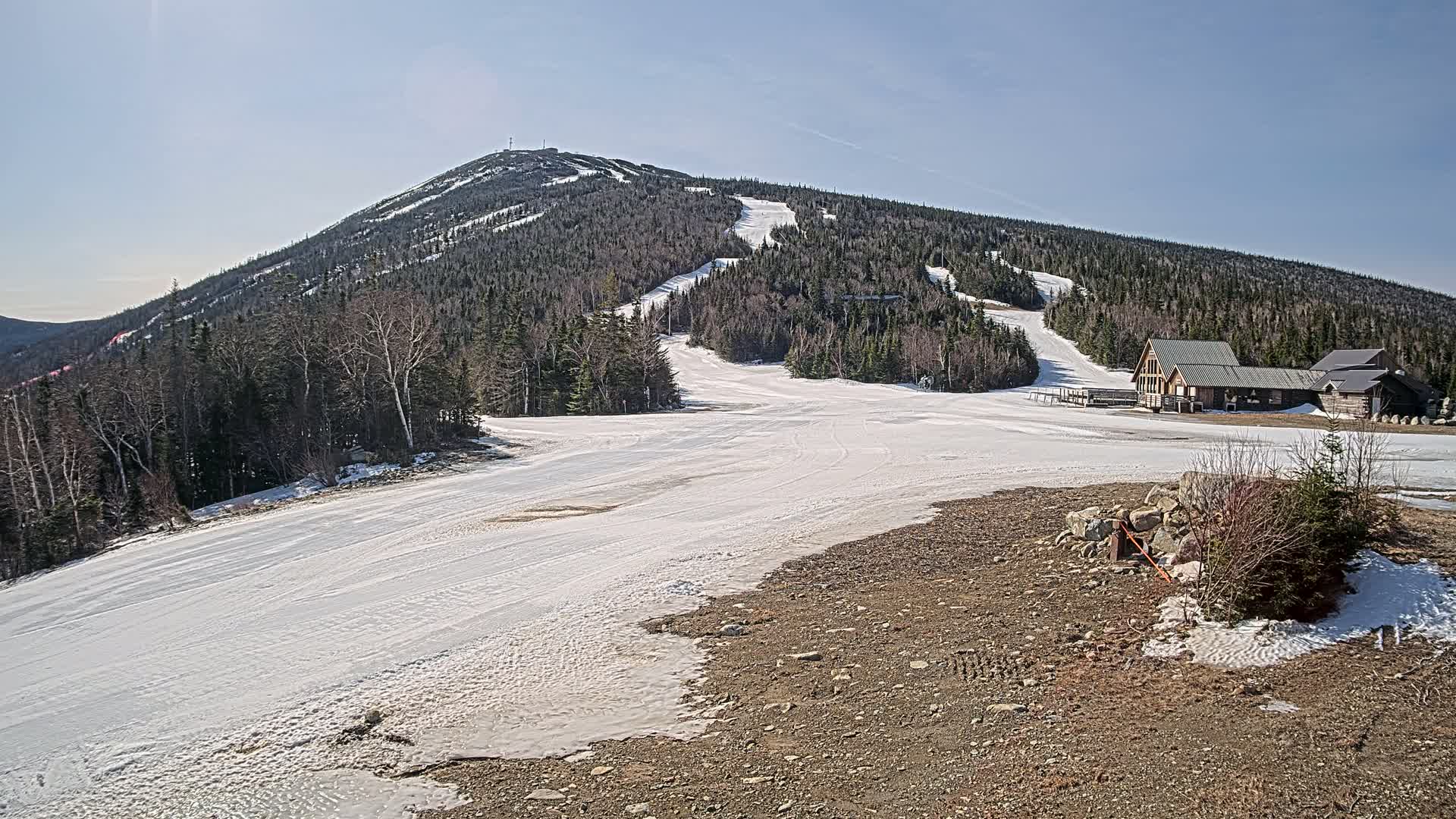 A snow-covered ski slope on a sunny day, with a small building at the base of a forested mountain.