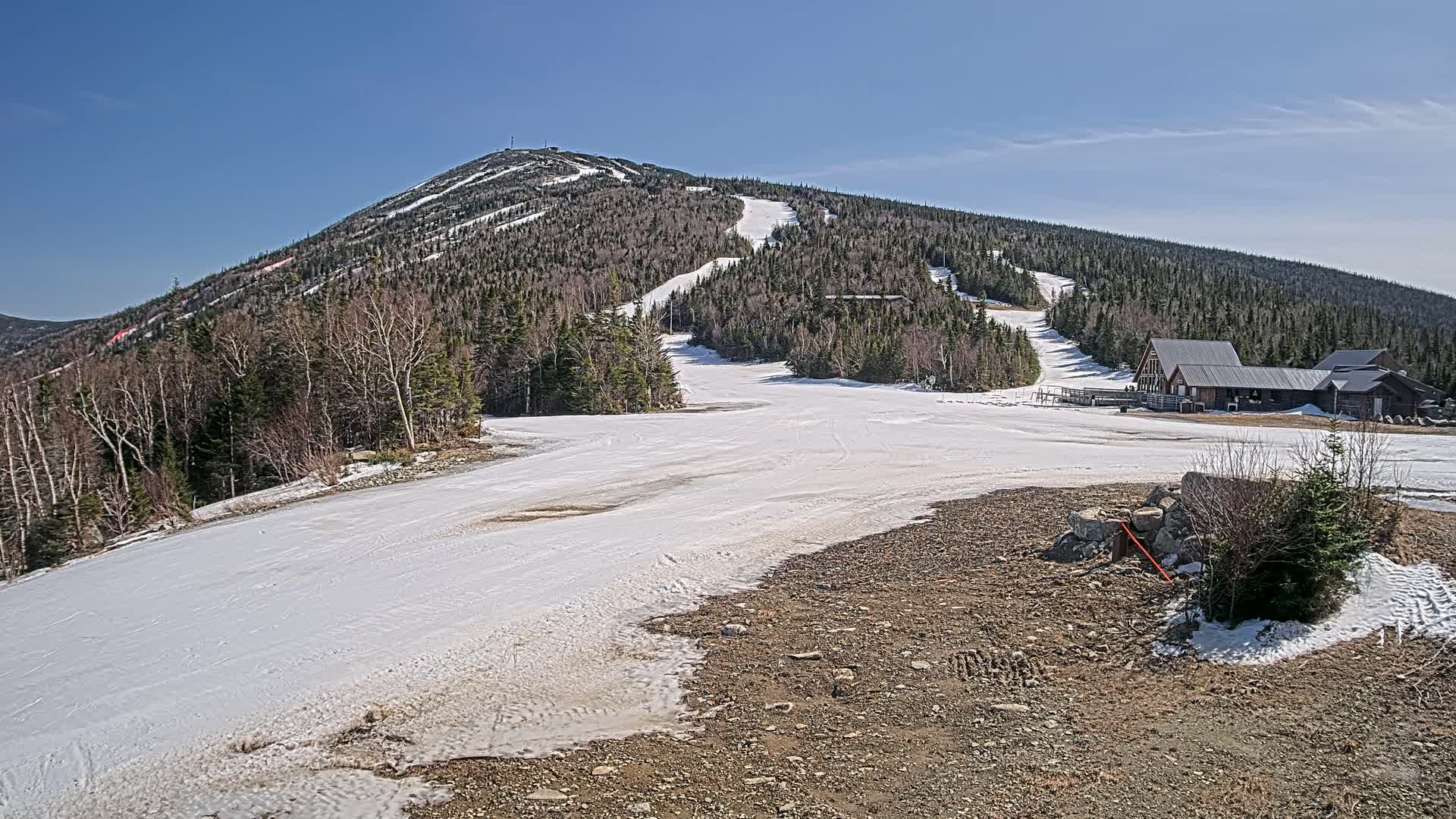 A snow-covered ski slope leads to a mountain peak under a clear blue sky, with a small building at the base of the slope.