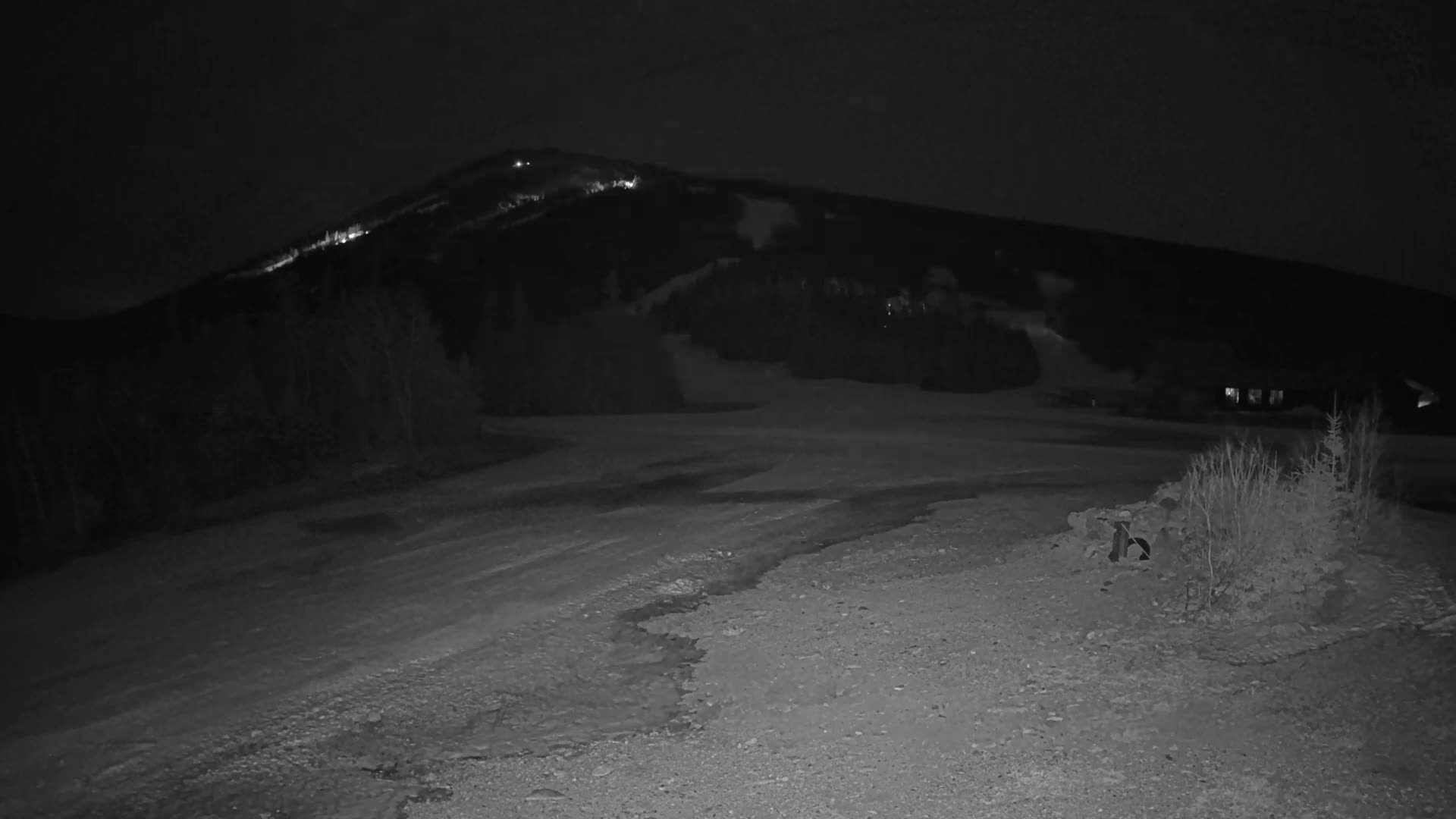 A snow-covered, dark, nighttime scene shows a sparsely lit mountain in the background and a gravel area in the foreground.