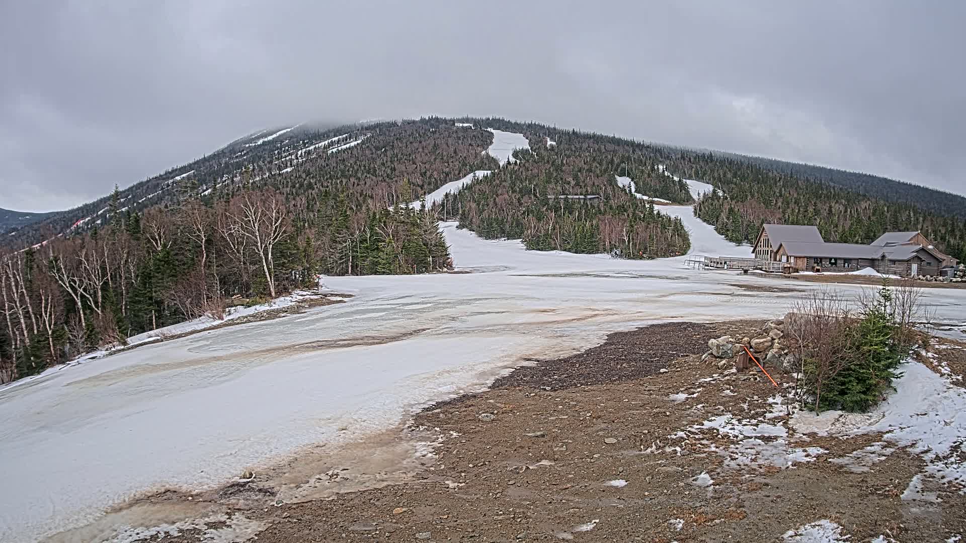 A mostly cloudy day reveals a snow-covered ski slope with a lodge at its base, partially melted snow, and patches of exposed ground.