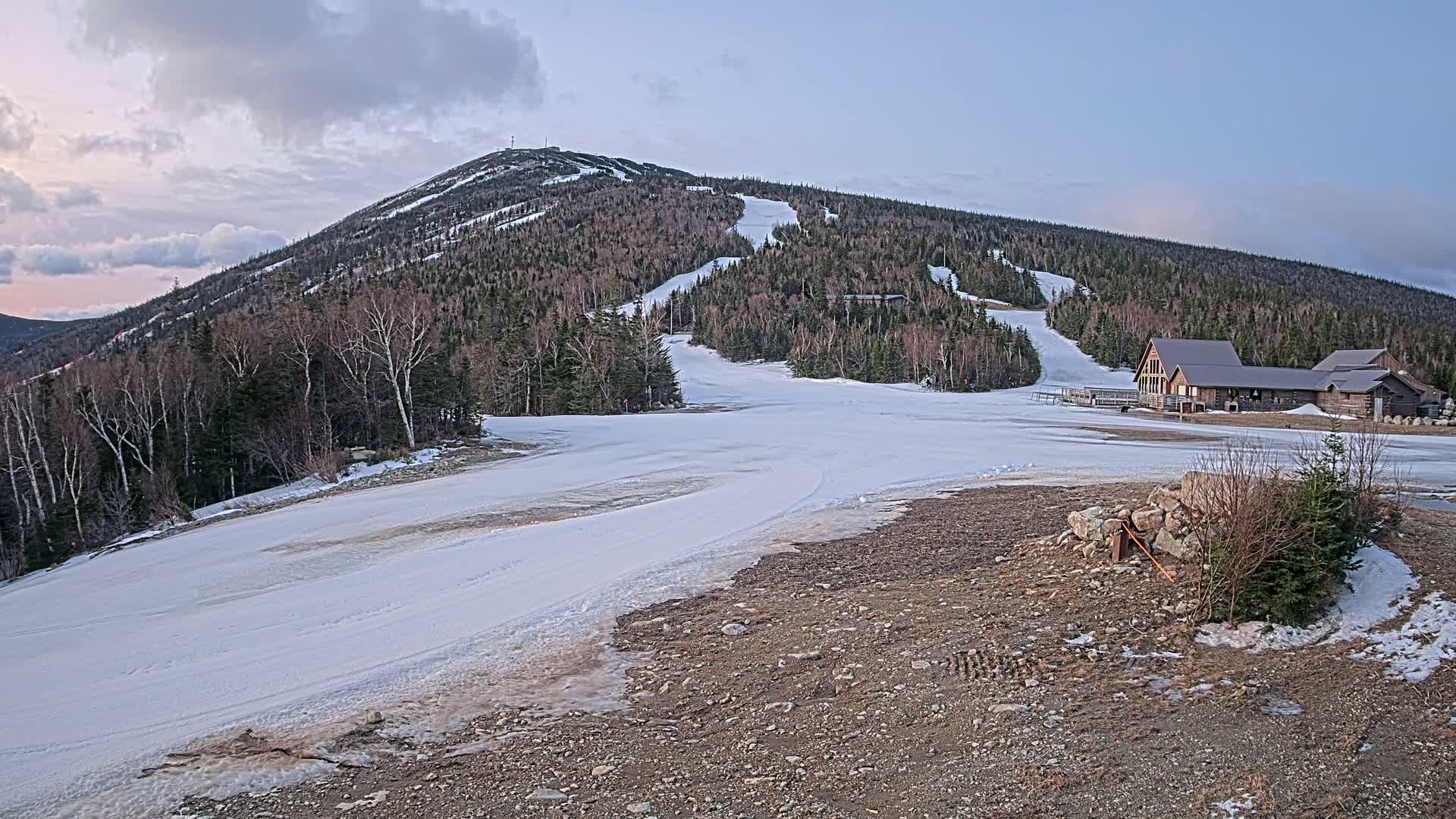 A snow-covered ski slope at the base of a mountain with a lodge, under a partly cloudy sky.