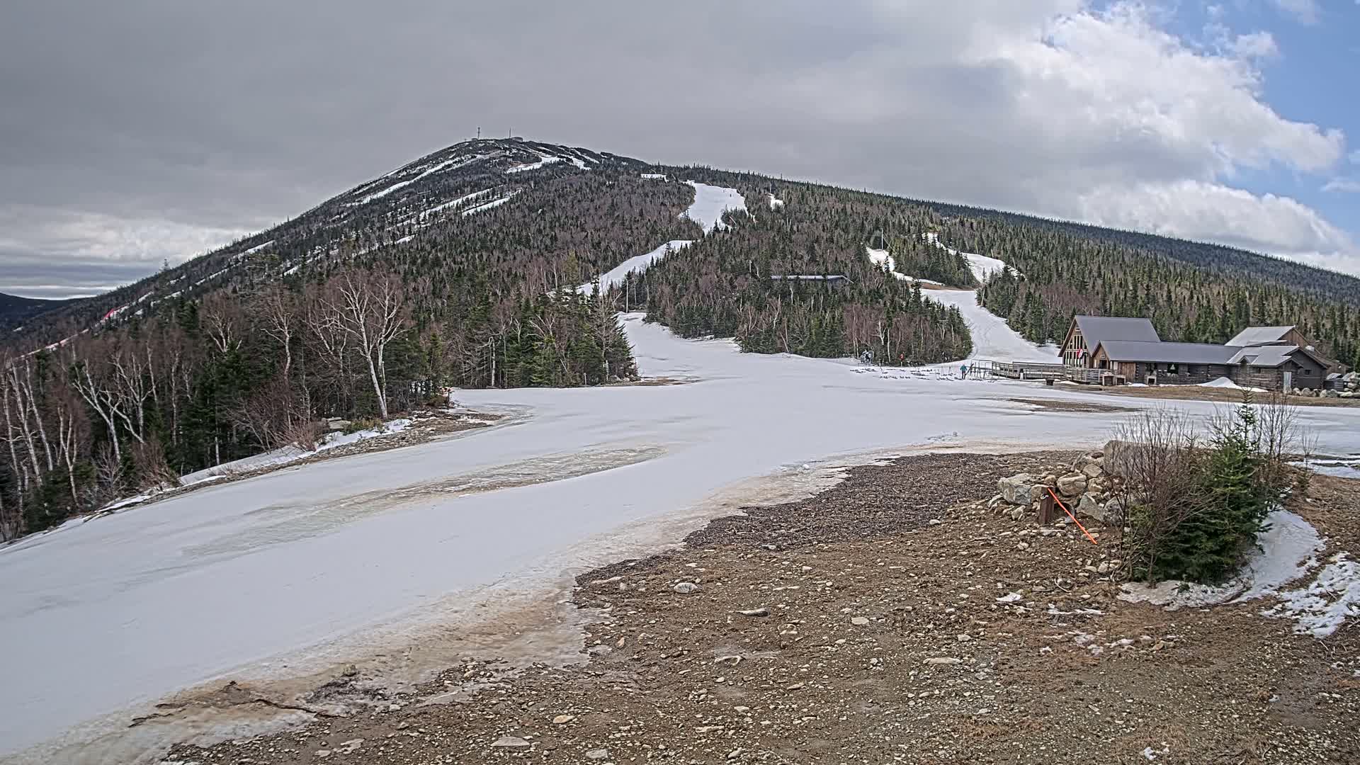 A snow-covered ski slope at the base of a forested mountain under a partly cloudy sky shows melting snow and a small building complex.