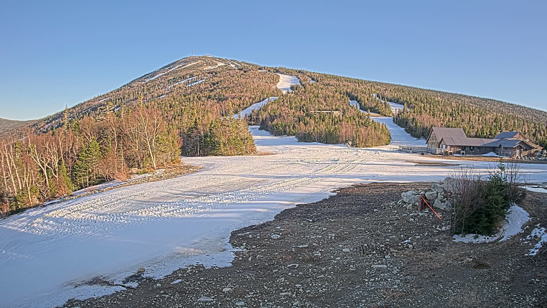A snow-covered ski slope at the base of a mountain, under a clear blue sky, with a building near the bottom of the slope.