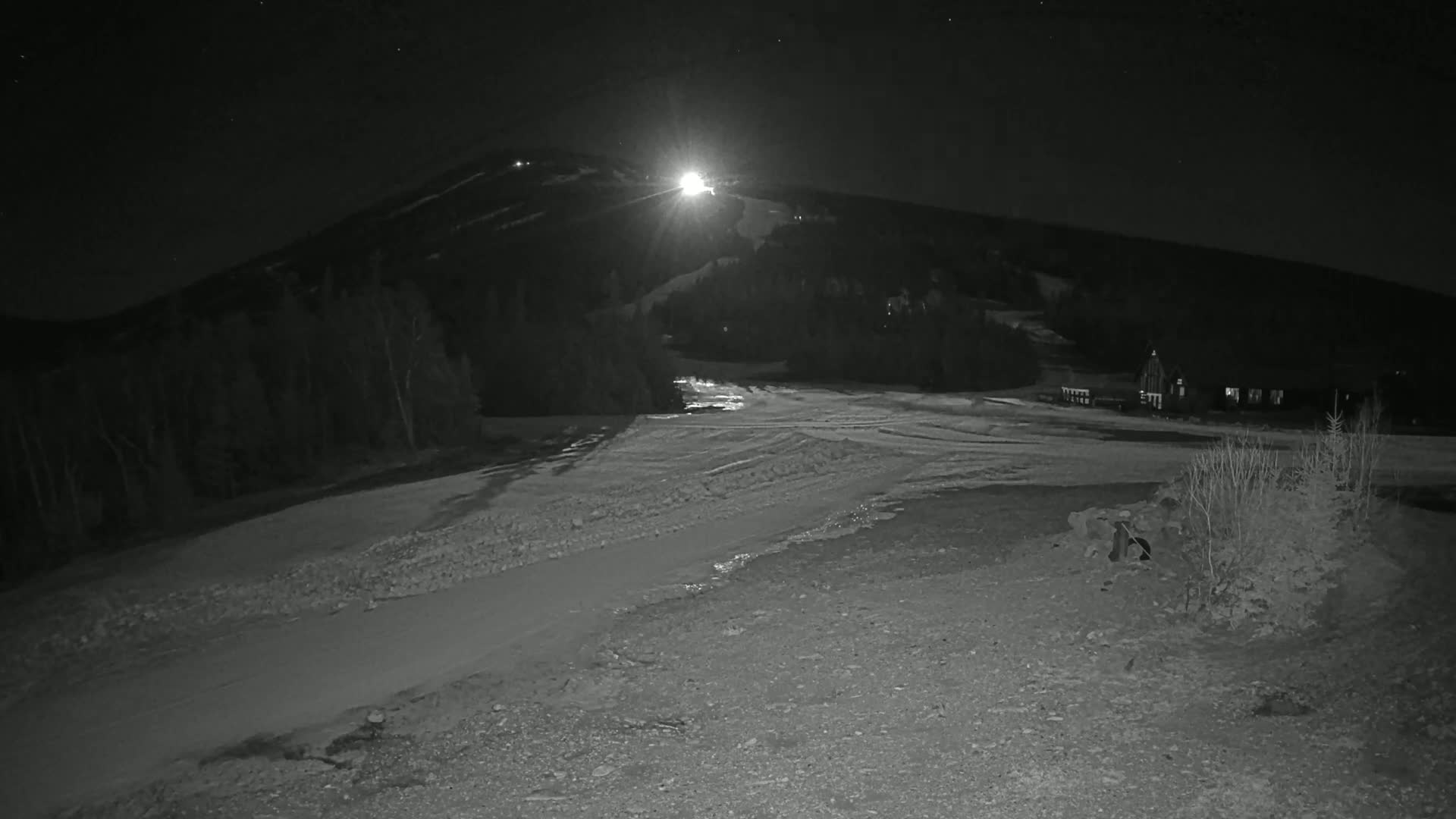 A snow-covered mountain slope is illuminated at night by a bright light, with a building visible in the lower right.