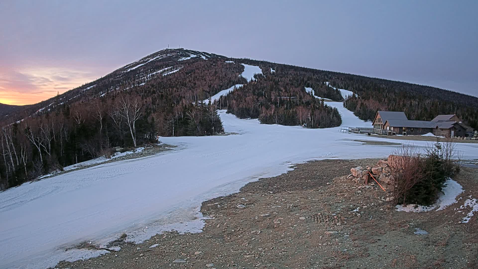 A snow-covered ski slope on a mountain at sunset, with a lodge at the base.