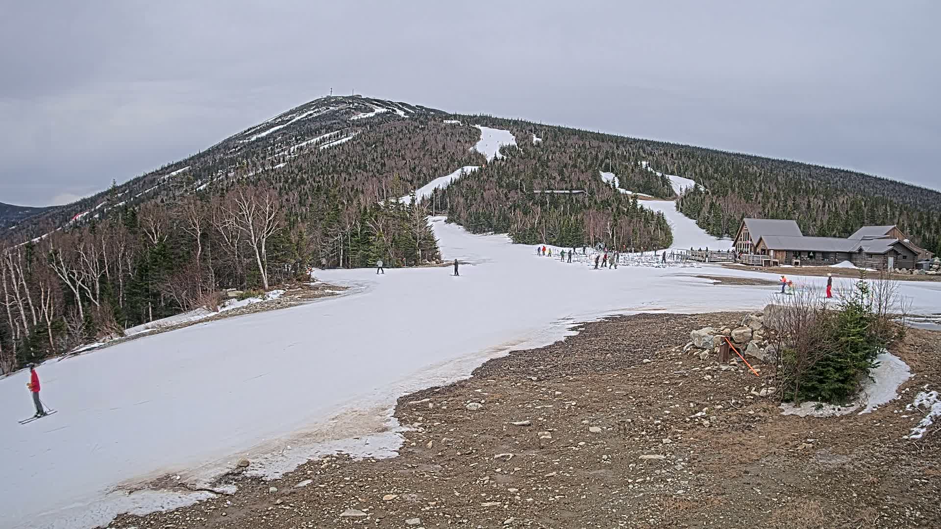 A snow-covered ski slope on a mostly cloudy day, with a few skiers visible and a wooden lodge at the base of a wooded mountain.